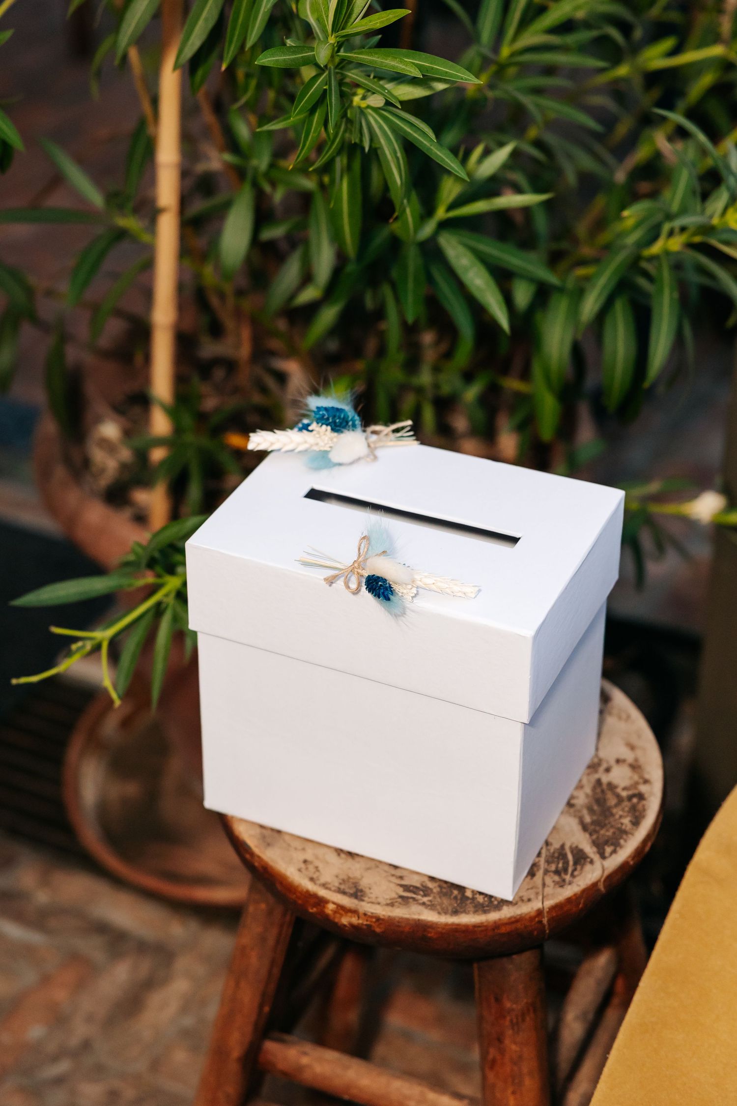 White wedding card box with blue floral decorations sitting on a wooden stool among tropical plants.