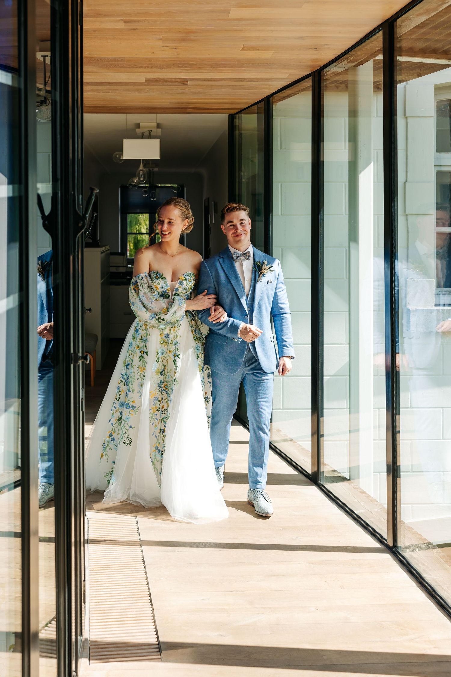 Bride in floral gown and groom in blue suit walking through glass doors at their wedding venue.