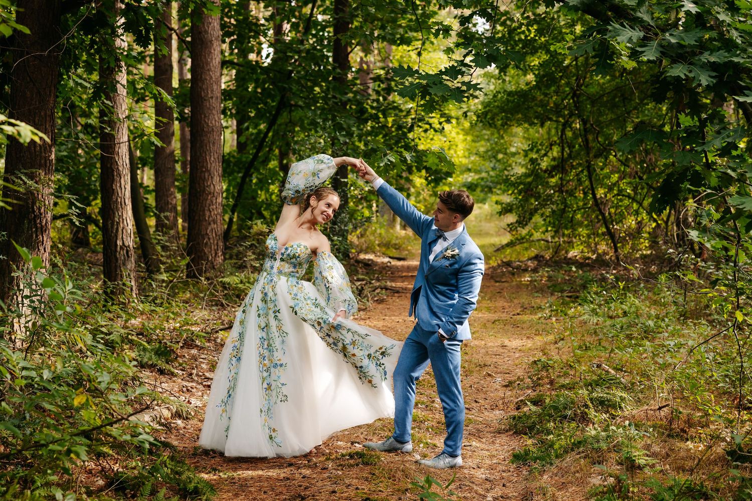Groom twirls bride in floral green wedding dress along a forest path in lush woodland setting.