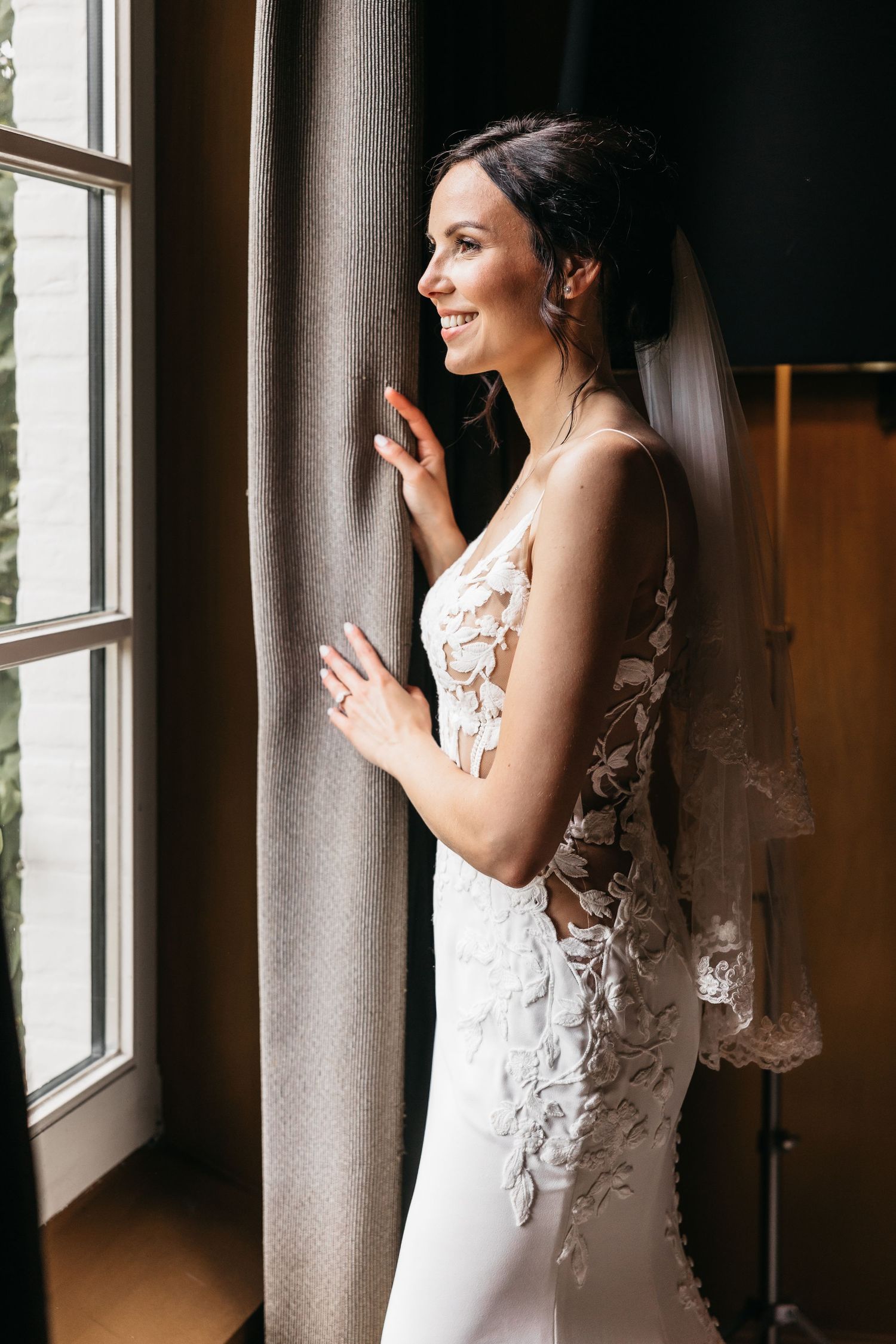 Smiling bride in lace wedding gown standing by a window holding sheer curtains.