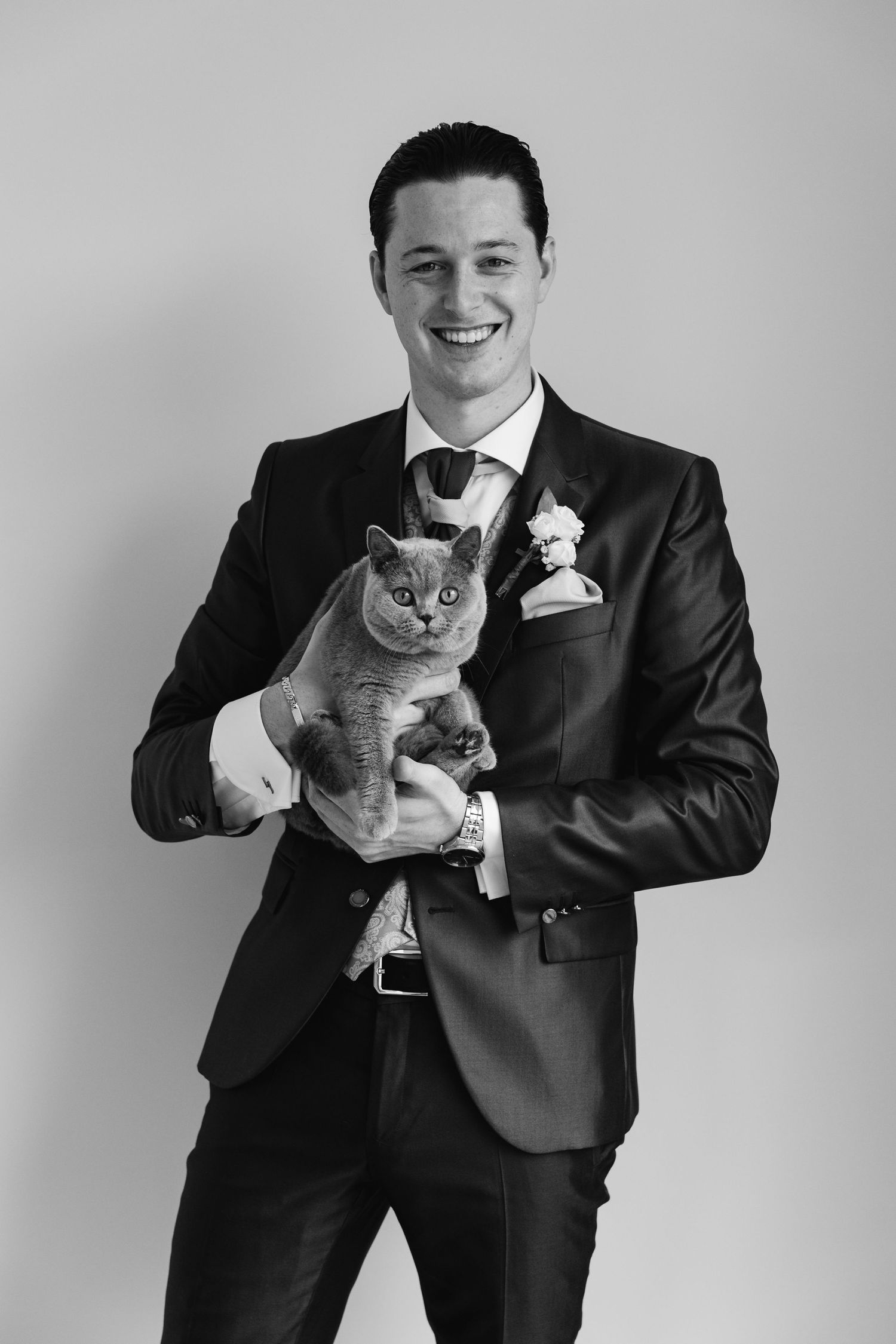 Black and white portrait of grinning groom in suit holding fluffy gray cat before wedding ceremony.