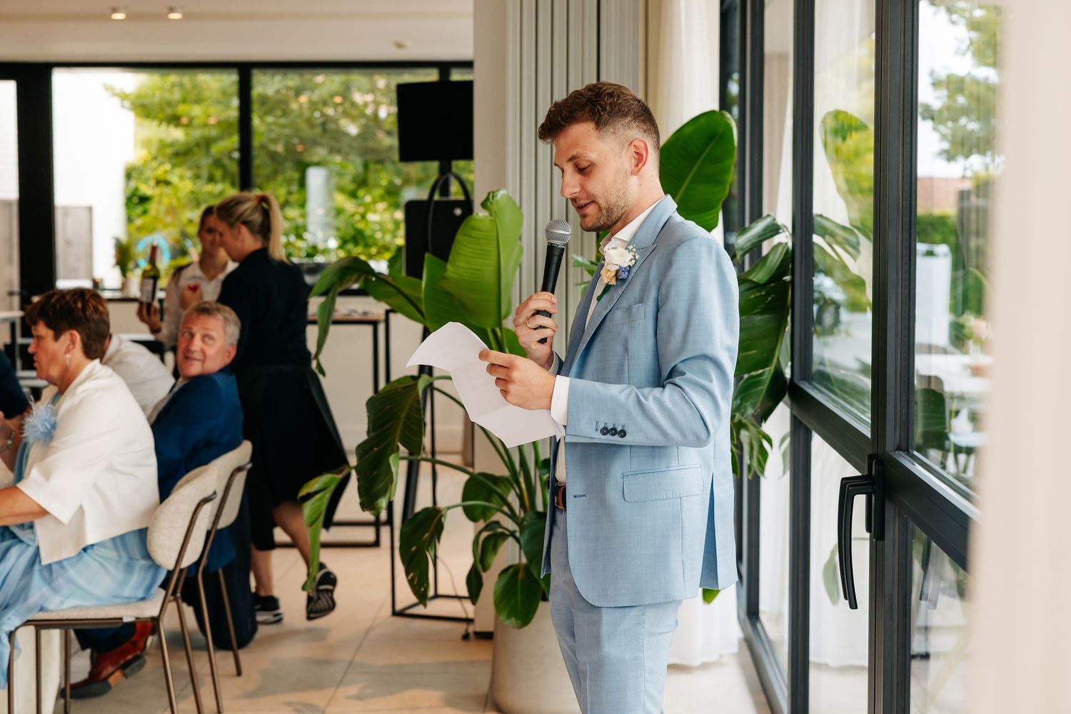 Man in light blue suit delivers speech with microphone at bright modern wedding reception venue.