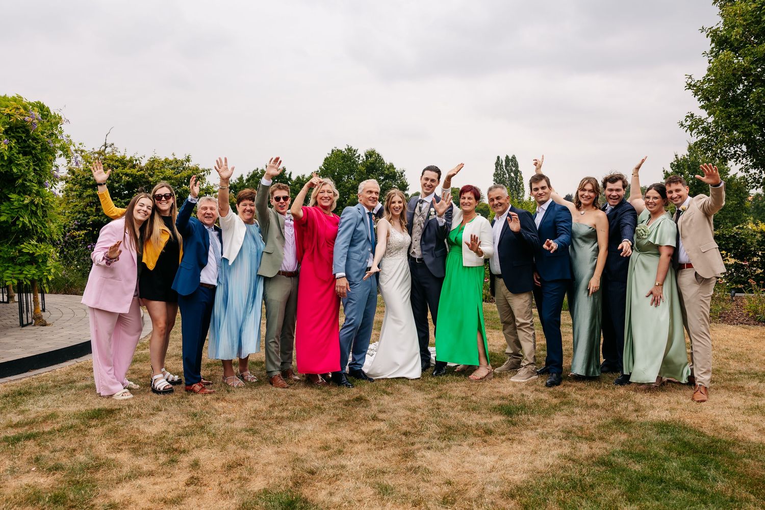 A cheerful wedding party group waves and smiles together on a grassy area at an outdoor celebration.
