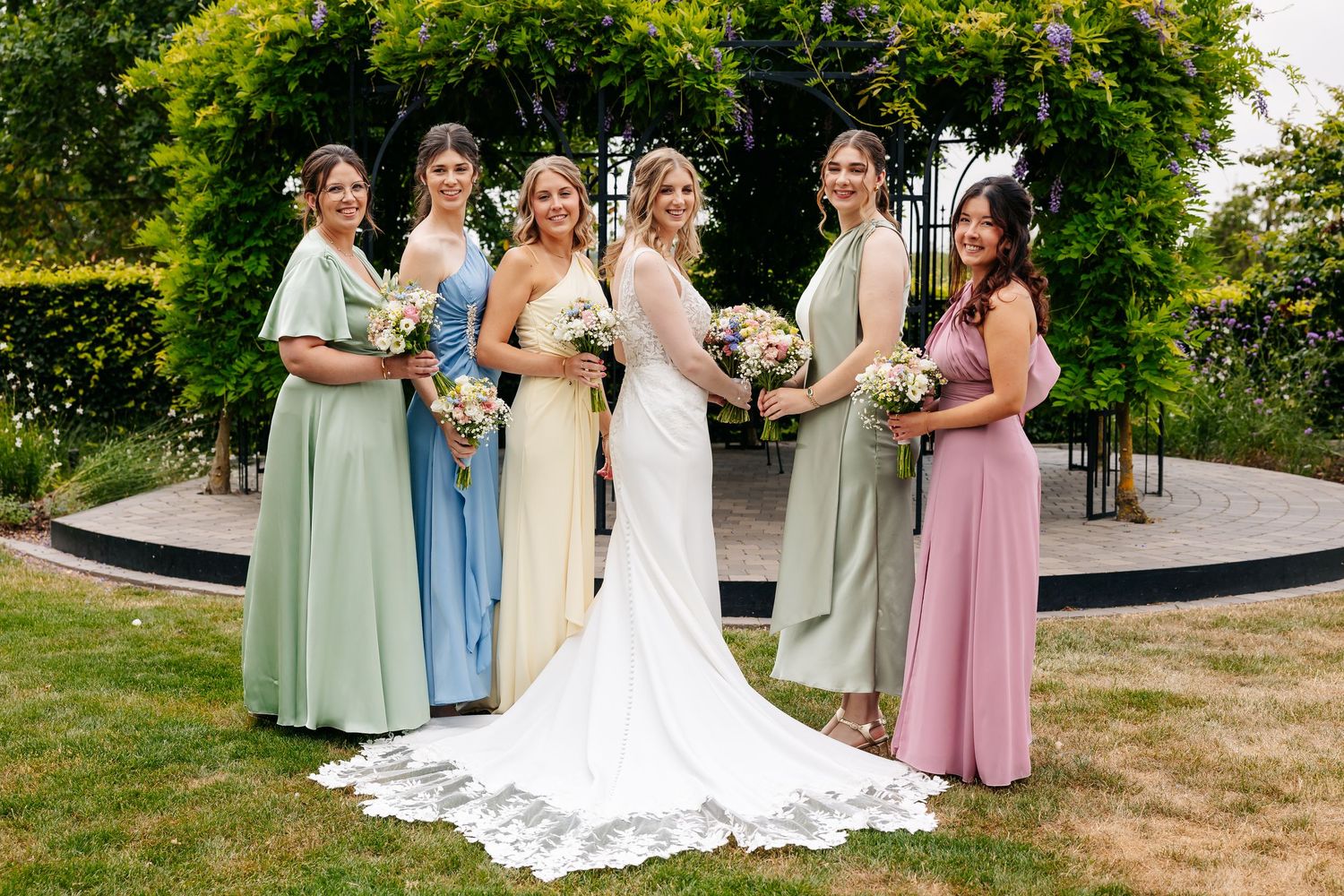 Bride and five bridesmaids in pastel mismatched gowns pose formally together in sunny garden setting.