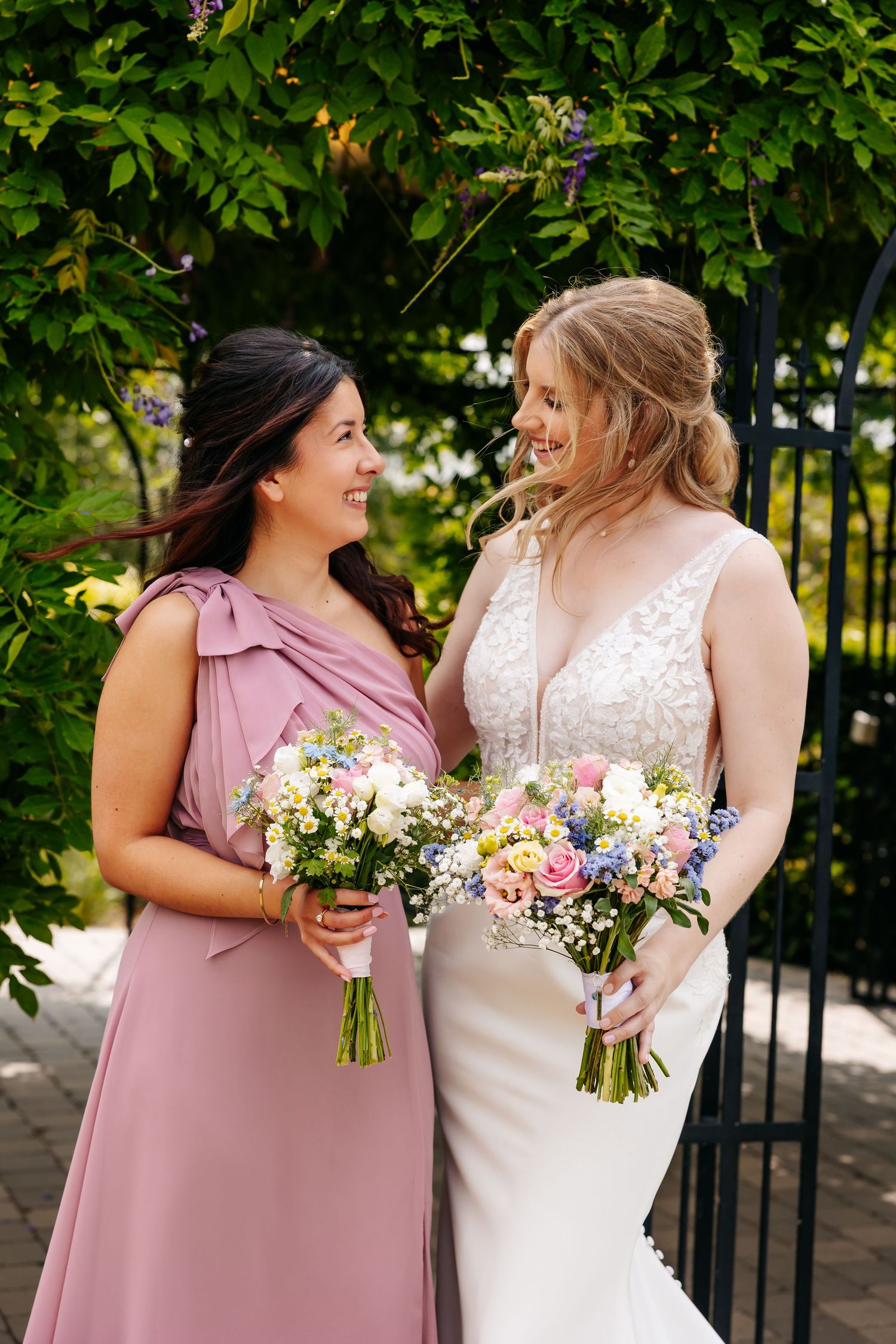 Bride in white lace gown and bridesmaid in pink dress share a smile while holding colorful floral bouquets.