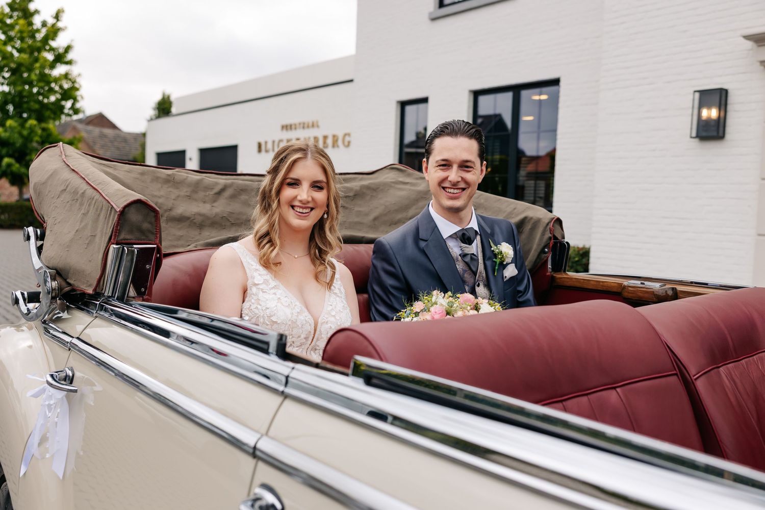 Happy newlyweds smiling in a classic vintage convertible with red leather interior at their wedding.