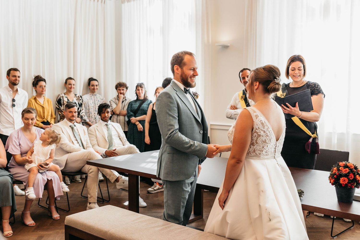 Bride and groom hold hands before officiant during indoor wedding ceremony with guests.