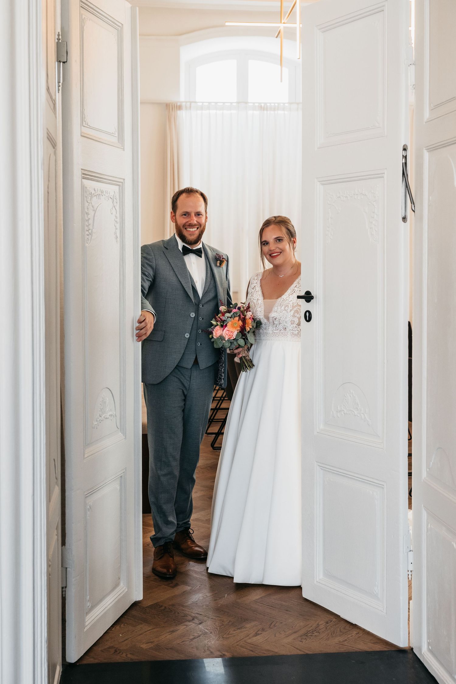 Smiling bride and groom standing in elegant white doorway, bride holding colorful bouquet, groom in gray suit with bow tie.