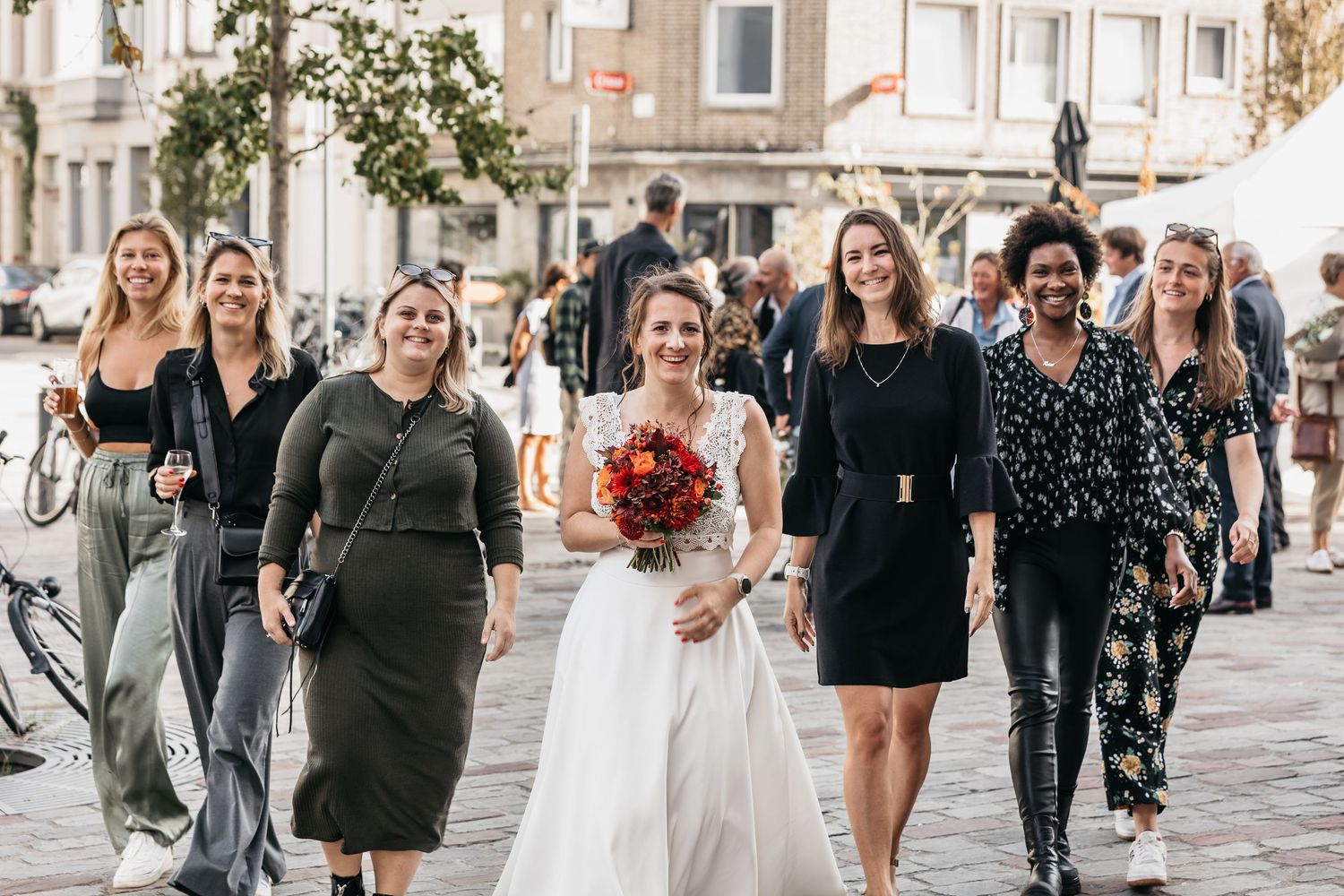Bride and bridesmaids walking together through a sunny cobblestone city square.