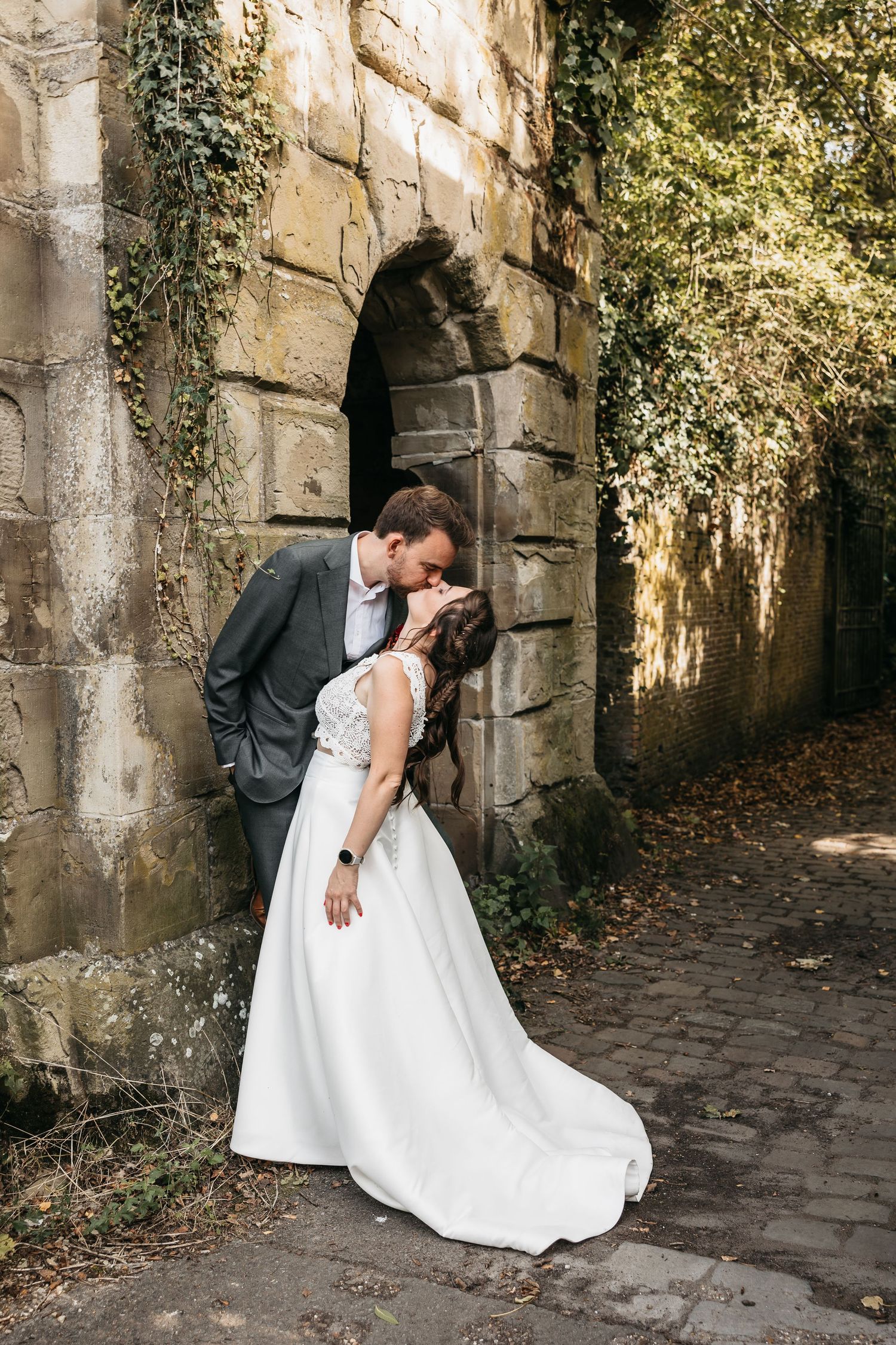 Bride and groom sharing a romantic kiss beside an ivy-covered stone archway on cobblestones.