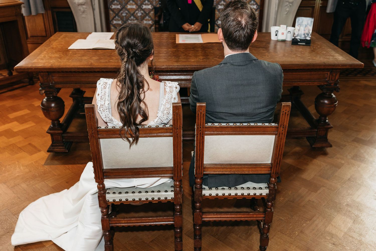 Bride and groom seated at ornate wooden table during civil wedding ceremony, viewed from behind.