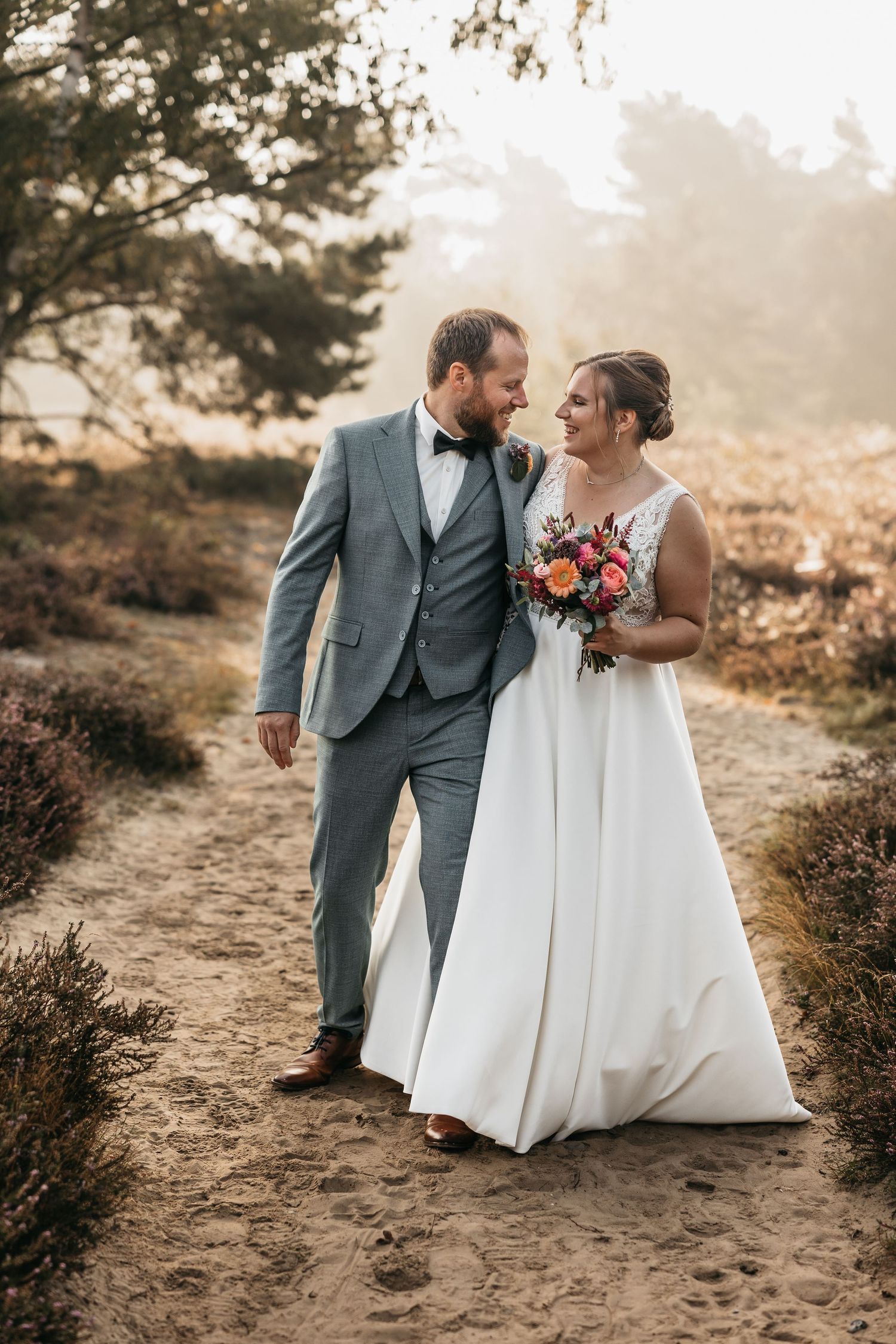 Bride and groom sharing a loving glance while walking together on a sandy heathland path in warm sunlight.