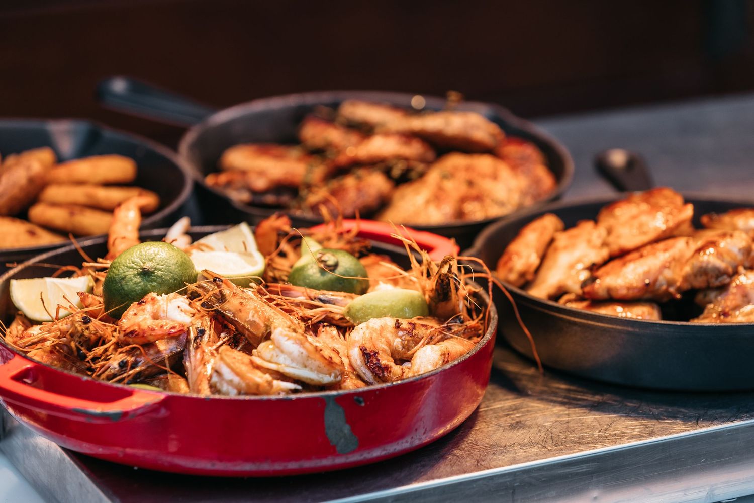 Cast iron pans filled with seasoned shrimp, crispy potatoes, and roasted chicken wings at a food station.