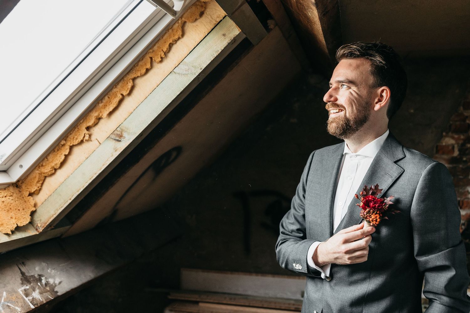 Smiling groom in gray suit with red boutonniere standing by skylight window in rustic loft.