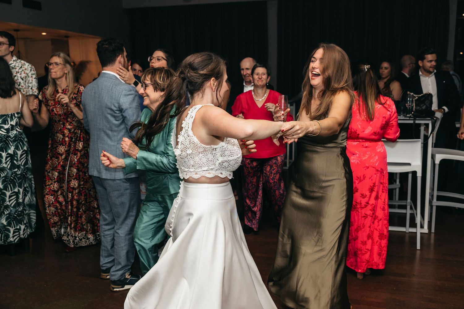 Bride in white gown dances joyfully with guests in colorful dresses at reception.
