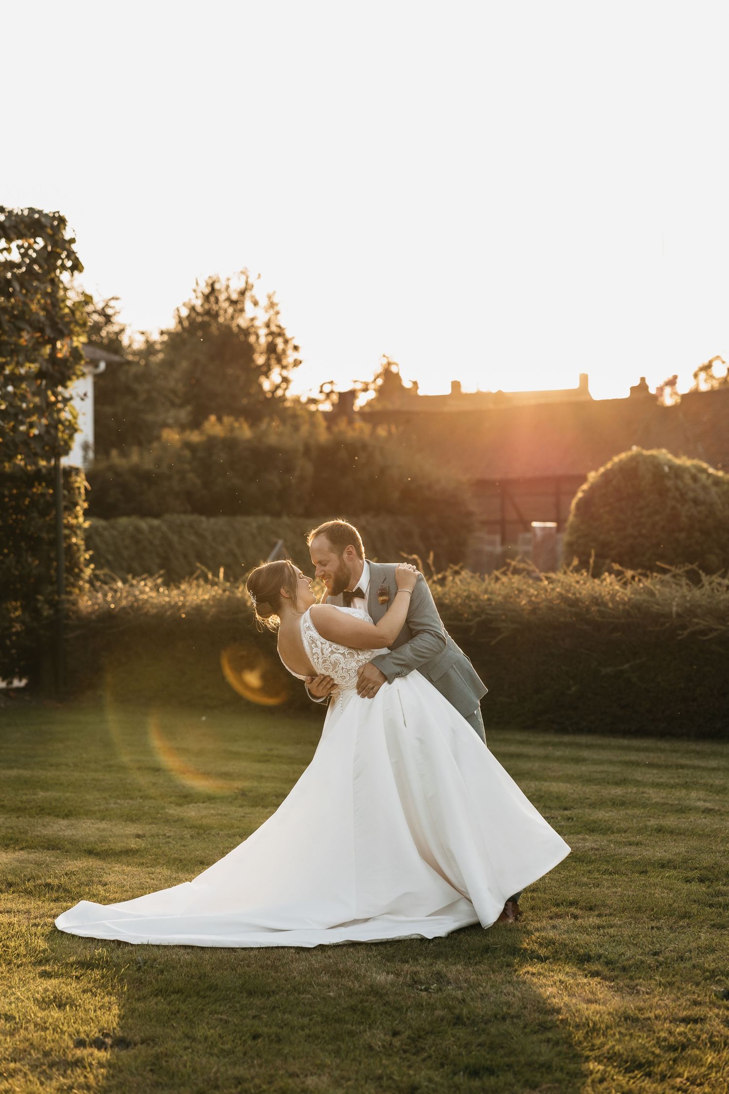 Bride and groom sharing a dip kiss on green lawn with glowing sunset backdrop.