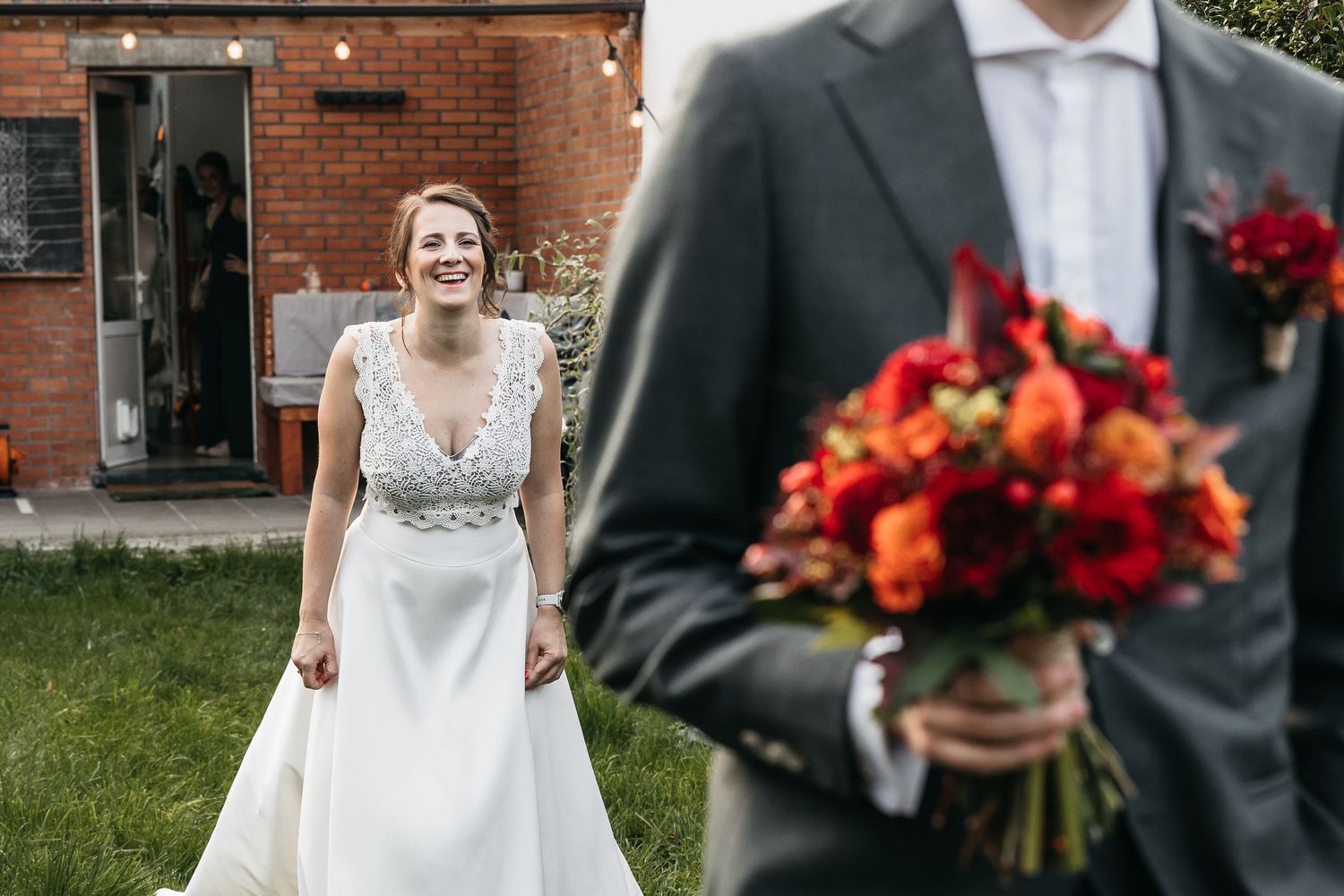 Bride laughing joyfully as groom holds a vibrant red and orange bouquet in the foreground.