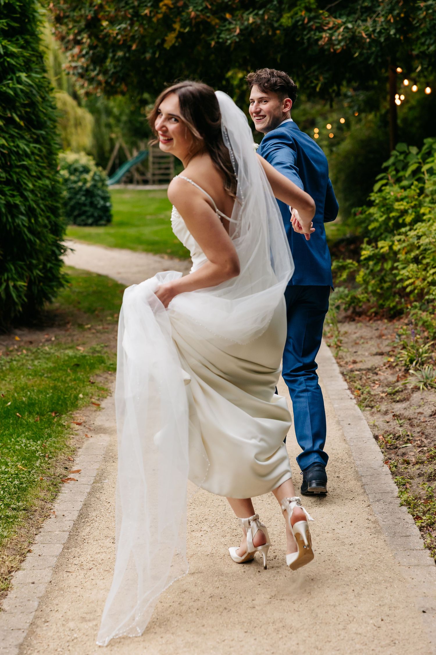 Bride and groom laughing and playfully walking along a garden path, bride lifting her white gown and veil.