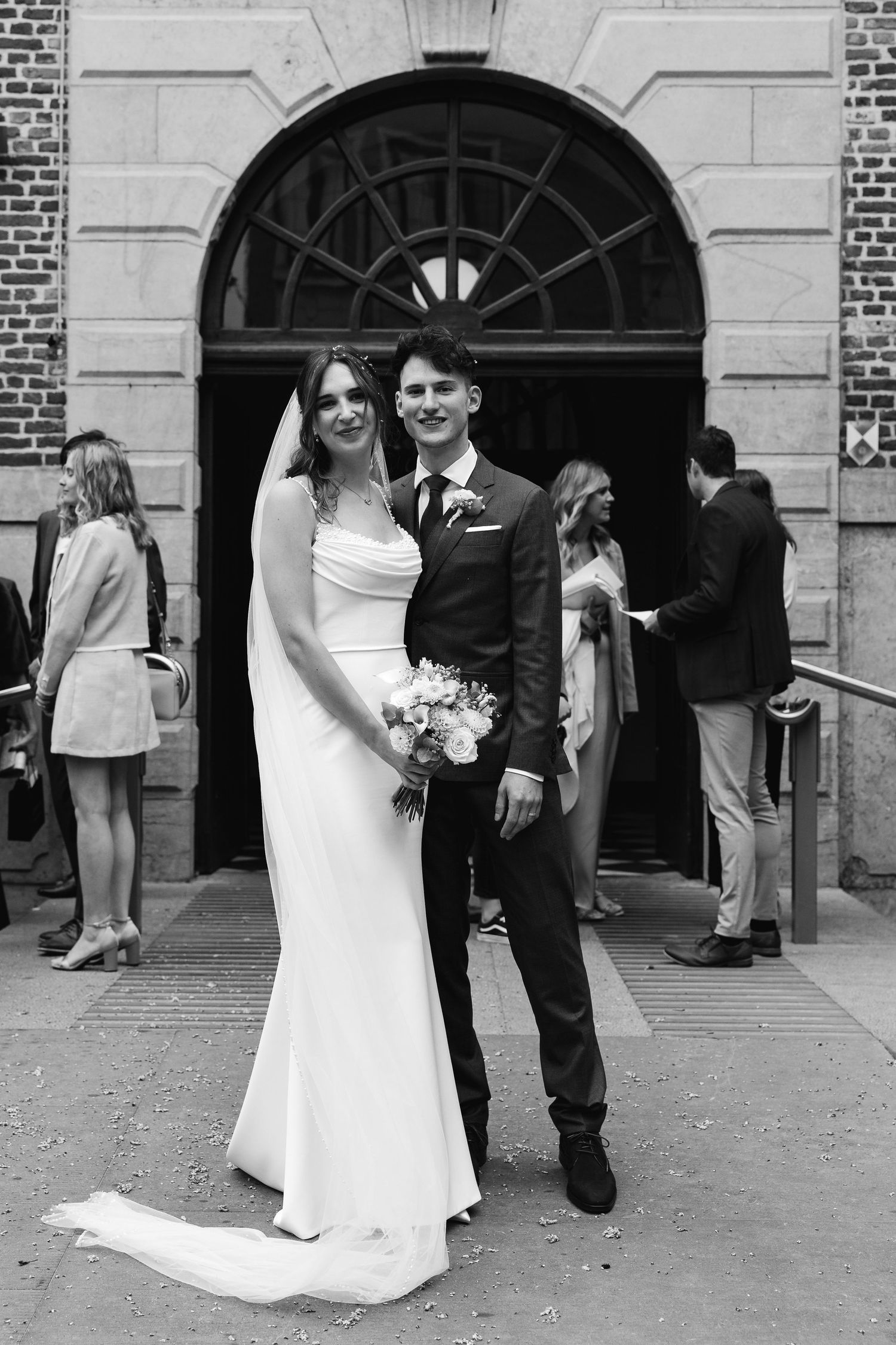 Black and white photo of newlyweds posing outside church, bride in white gown, groom in dark suit.