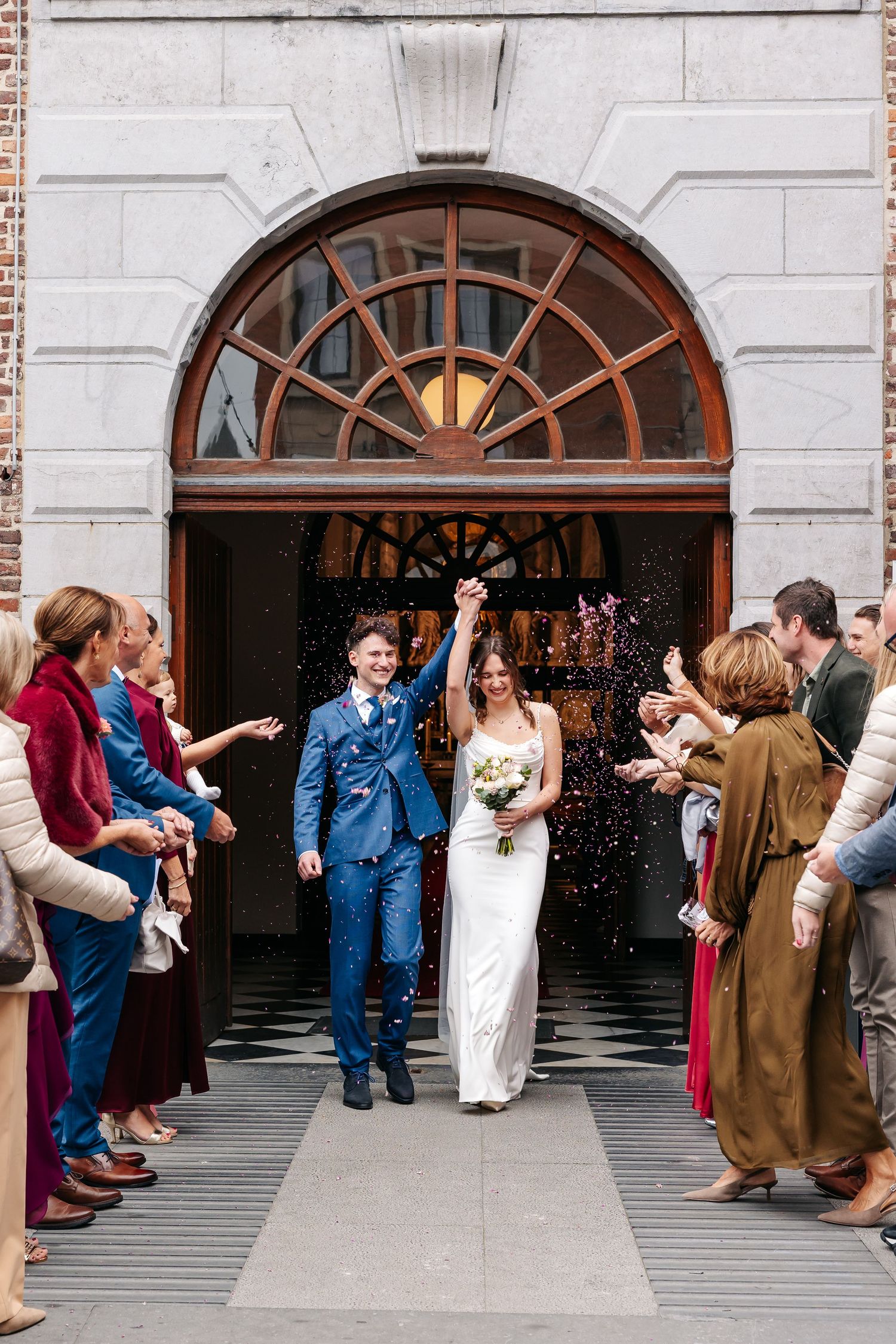 Joyful newlyweds exit church in confetti shower, groom in blue suit, bride in white gown.