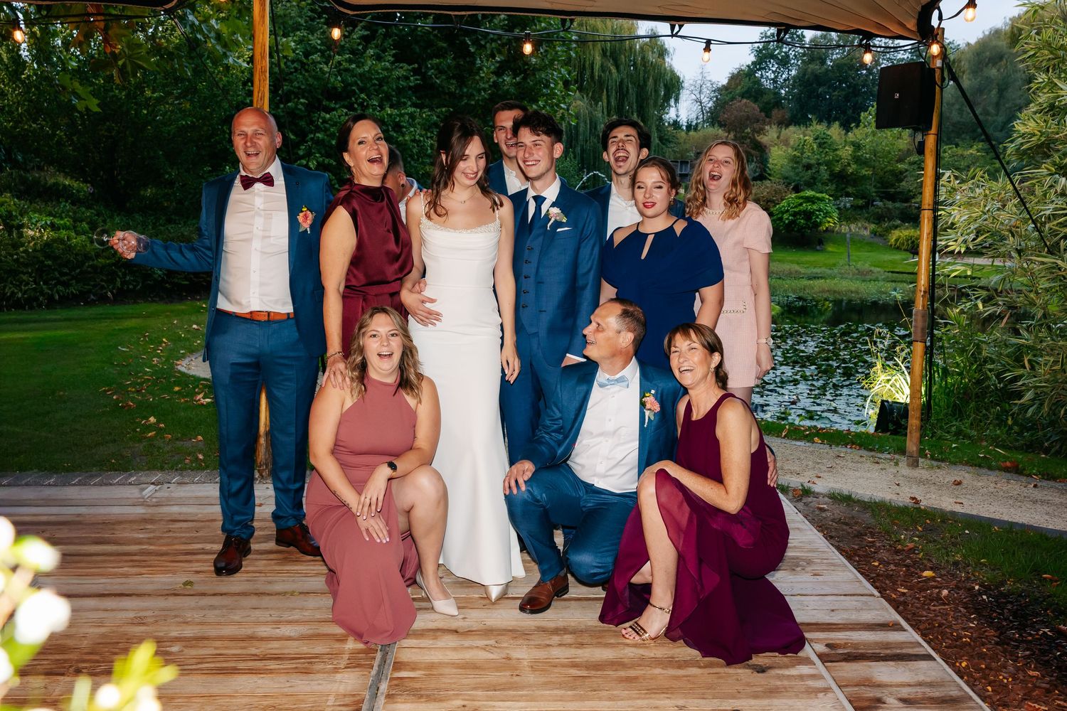 Wedding party group photo on wooden deck surrounded by lush greenery near a garden pond.
