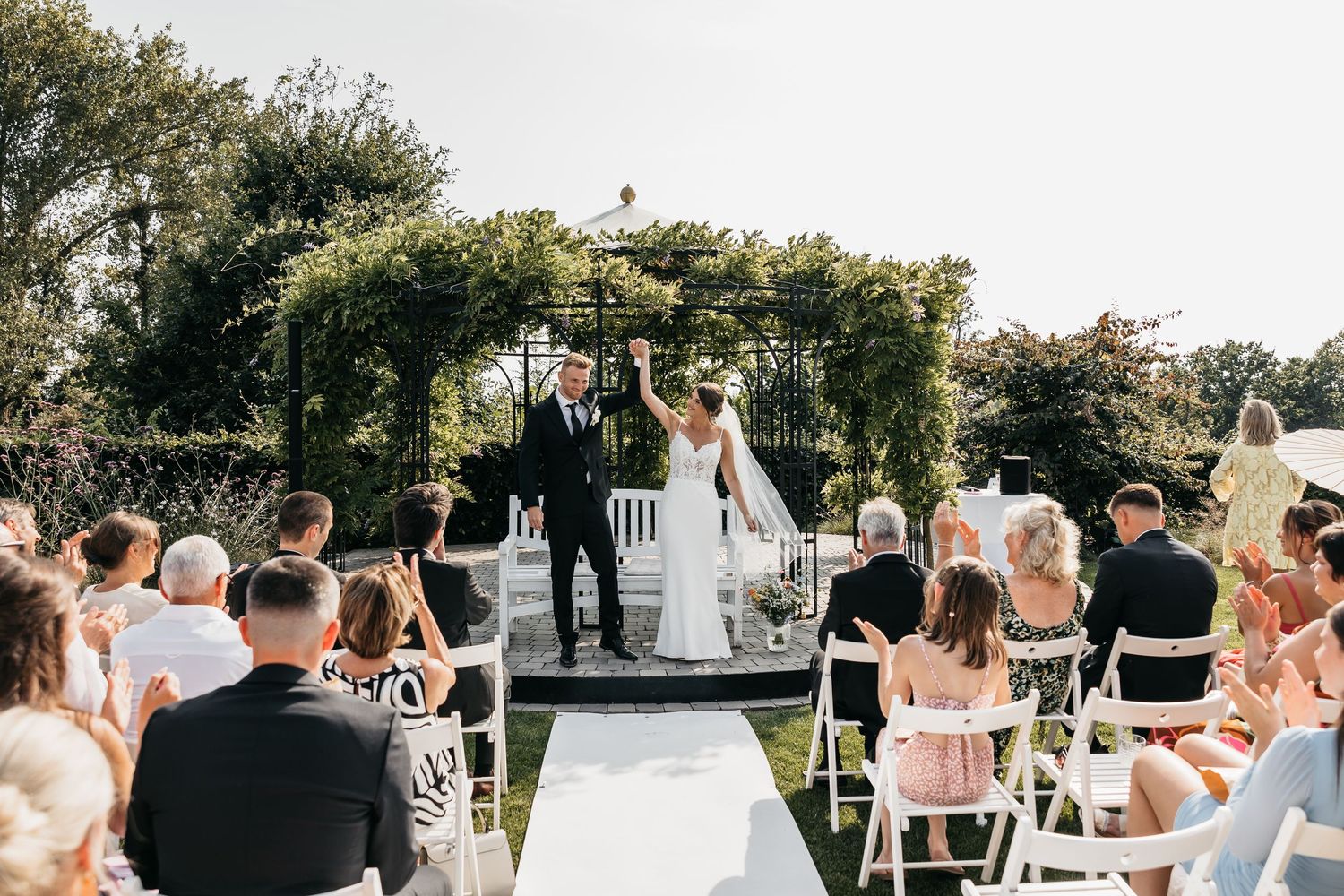 Bride and groom raise hands in celebration at outdoor garden wedding ceremony with seated guests.