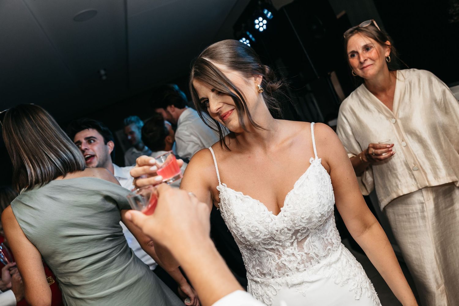 Bride in lace wedding gown accepts a drink from guest during lively wedding reception.