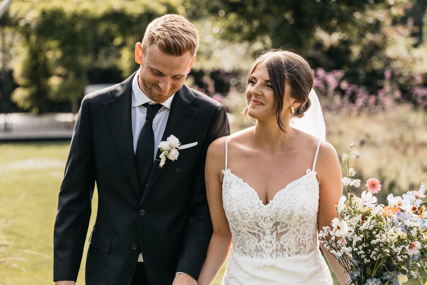 Bride in lace gown and groom in black suit share a tender glance in a garden setting.