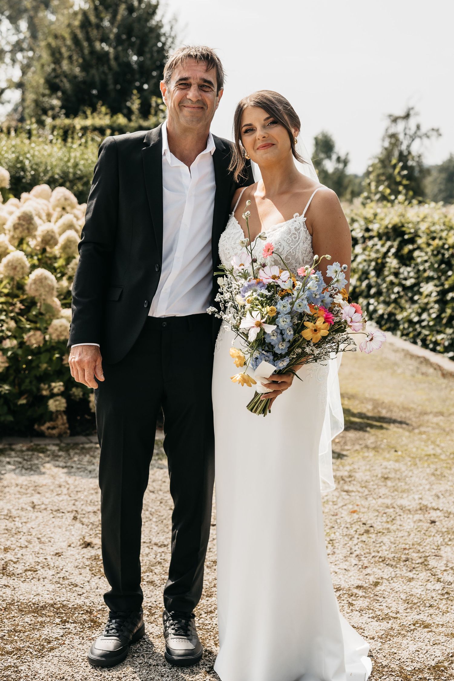 Bride in white lace gown holding colorful bouquet posing with man in black suit in garden setting.