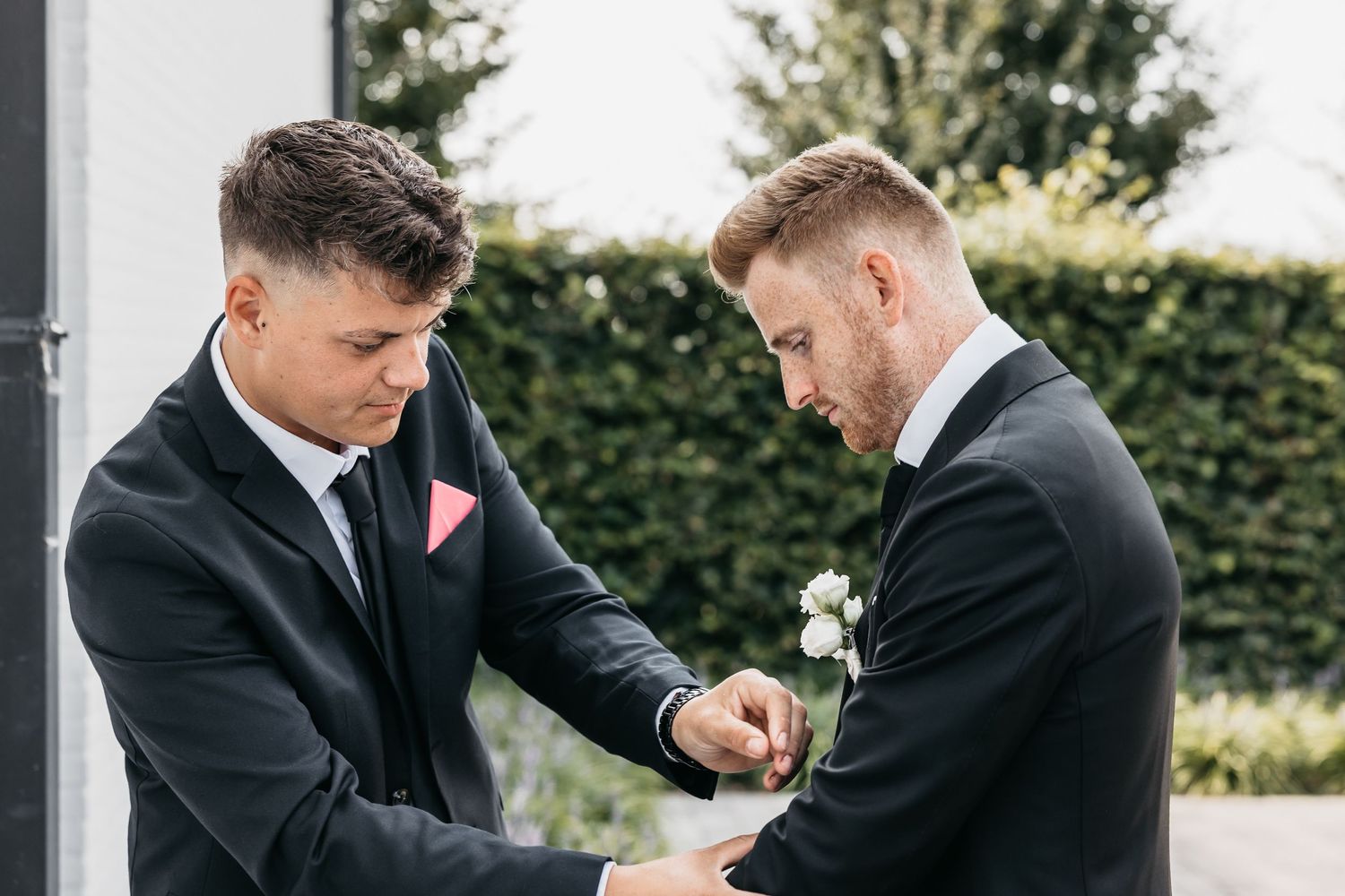 A groomsman pins a white boutonniere onto another mans black suit lapel outdoors.
