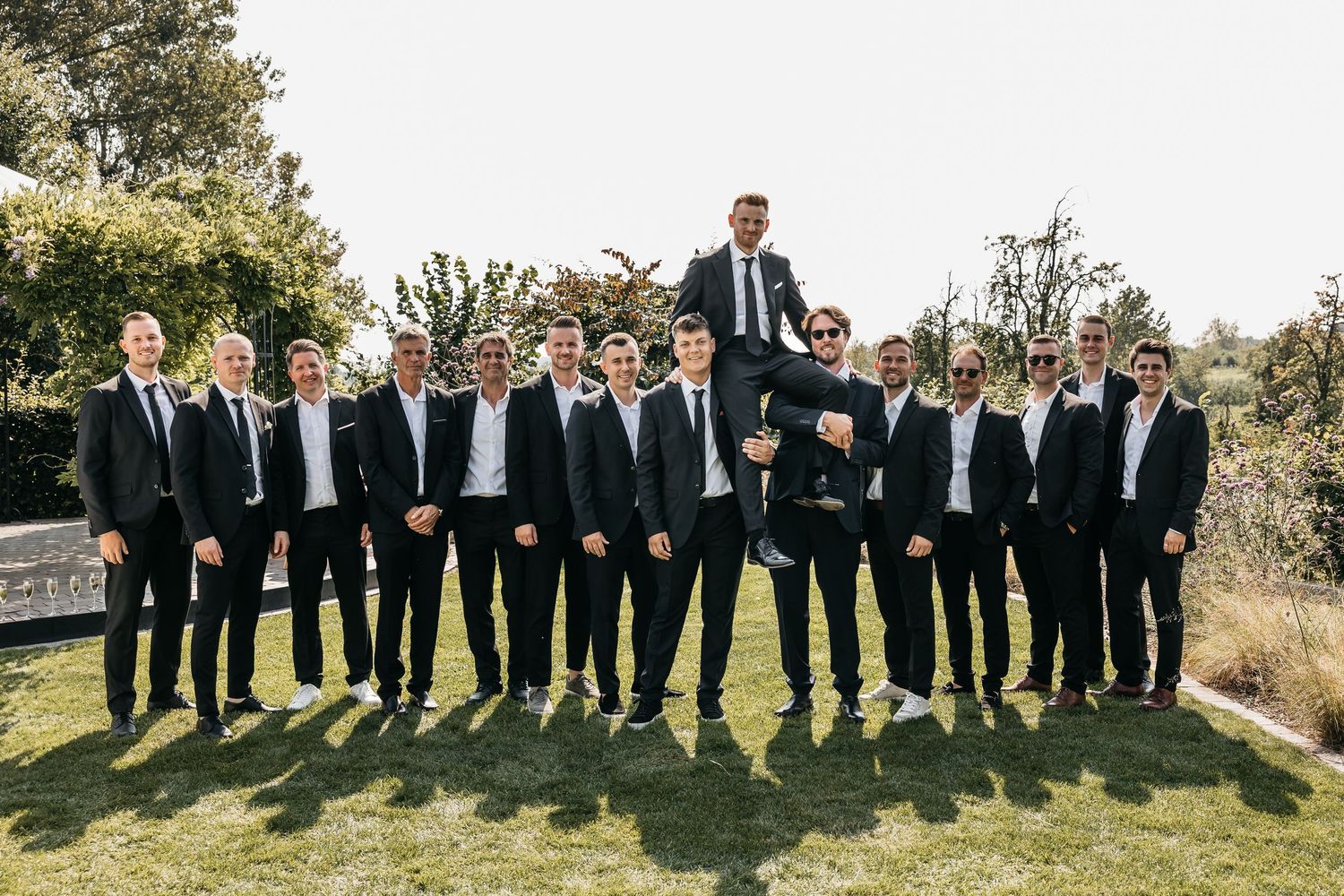 Large group of groomsmen posing with groom elevated on shoulders in sunny garden.