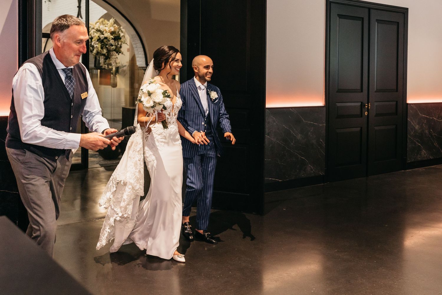 Bride in lace gown and groom in blue suit make grand entrance at wedding reception with escort.