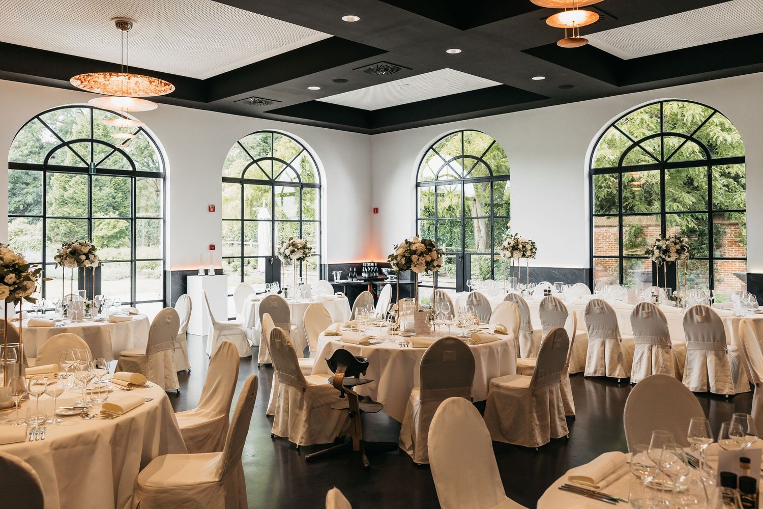 Bright wedding reception venue with large arched windows, round tables, white chair covers, and floral decor.