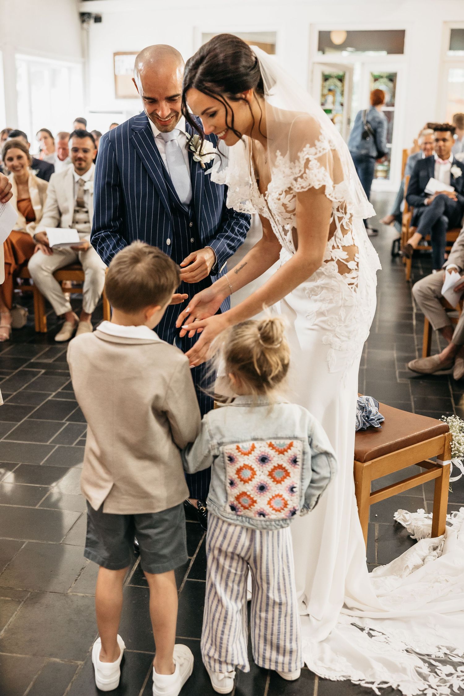 Smiling bride and groom interacting with flower girl and ring bearer during indoor wedding ceremony.