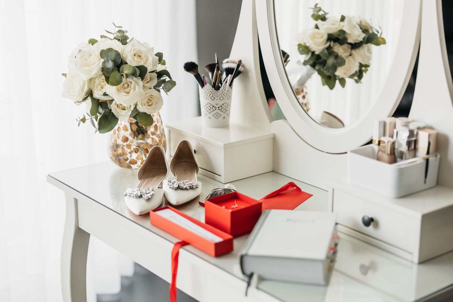 Bridal vanity table with white heels, red gift box, roses, and makeup accessories.