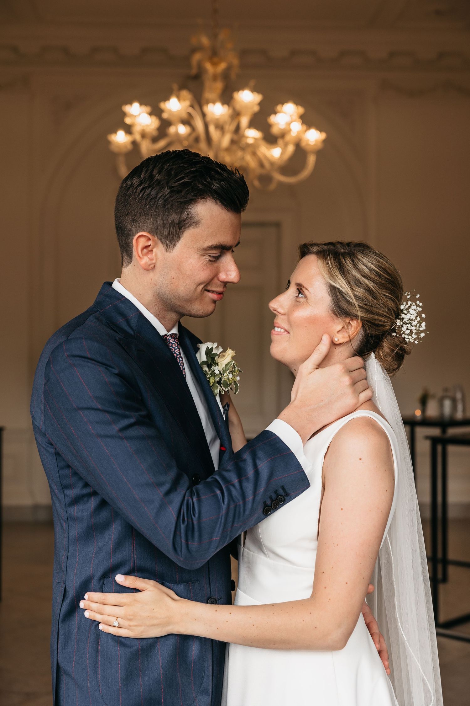 Groom tenderly cups brides face as they gaze at each other beneath a glowing chandelier indoors.