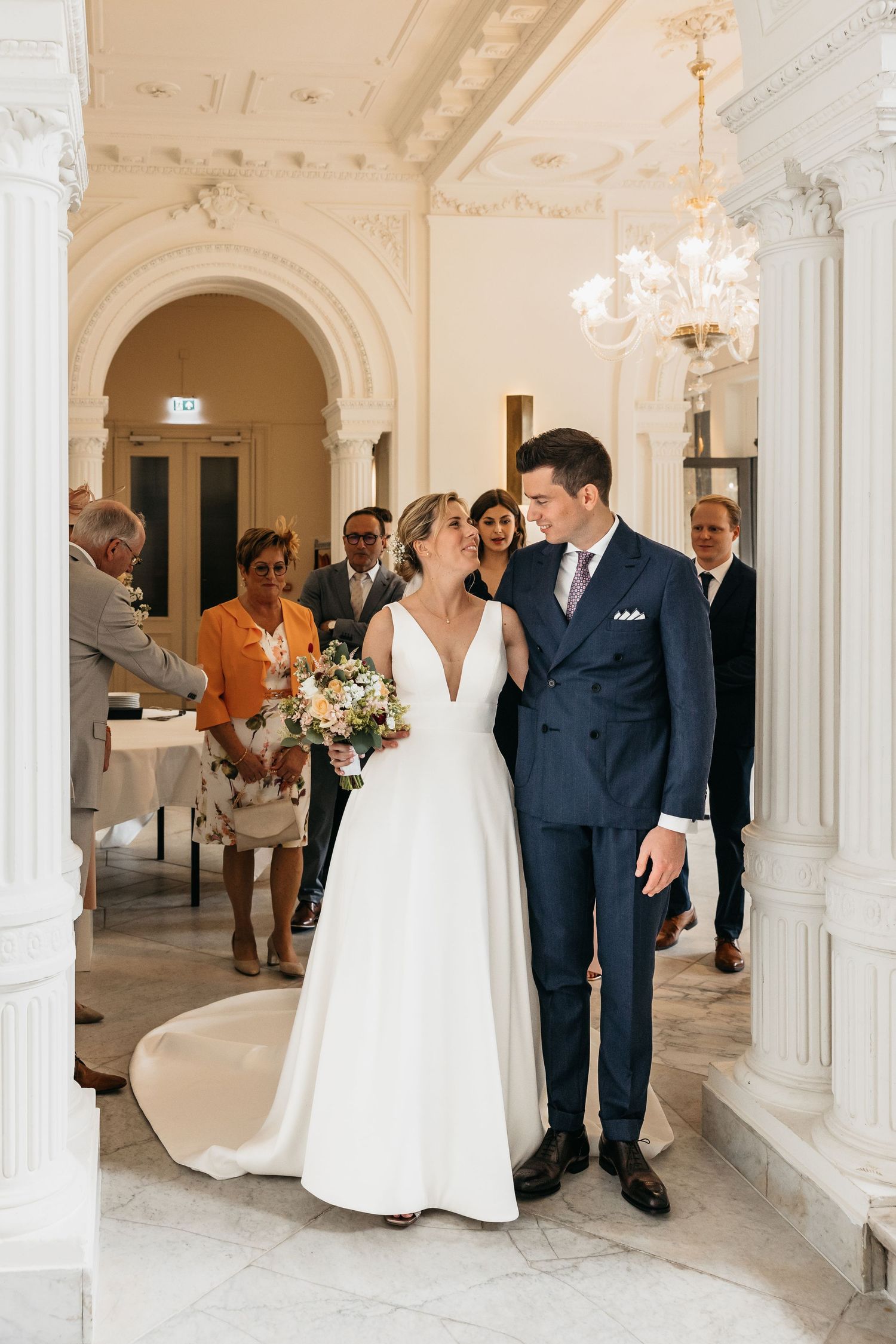 Newlyweds walk together through elegant white arched hallway with wedding guests.
