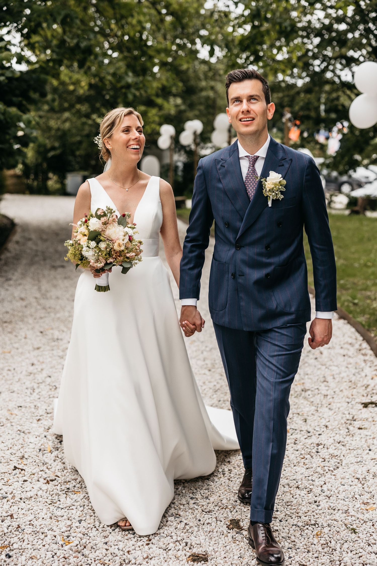Laughing bride and groom stroll hand in hand along gravel path at elegant estate wedding.