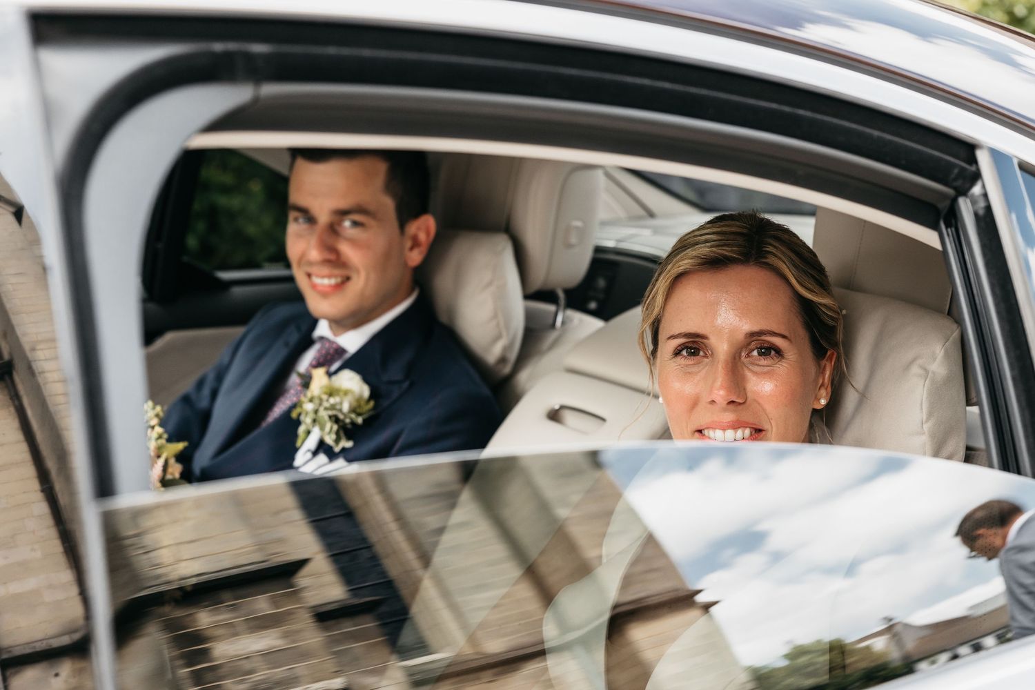 Smiling bride and groom seated in a luxury wedding car viewed through the window.