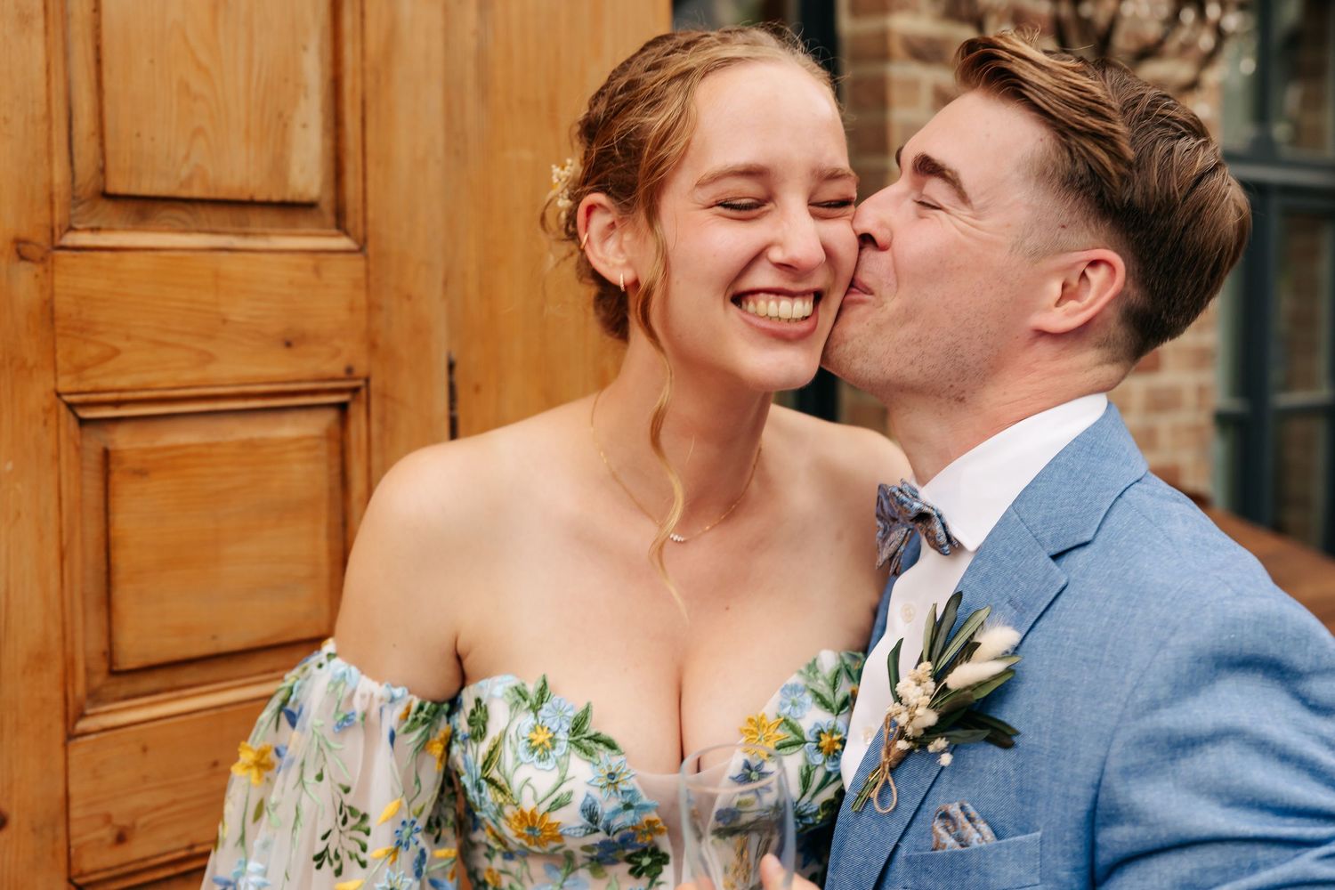 Groom in blue suit kissing bride on cheek as she smiles, standing before rustic wooden door.