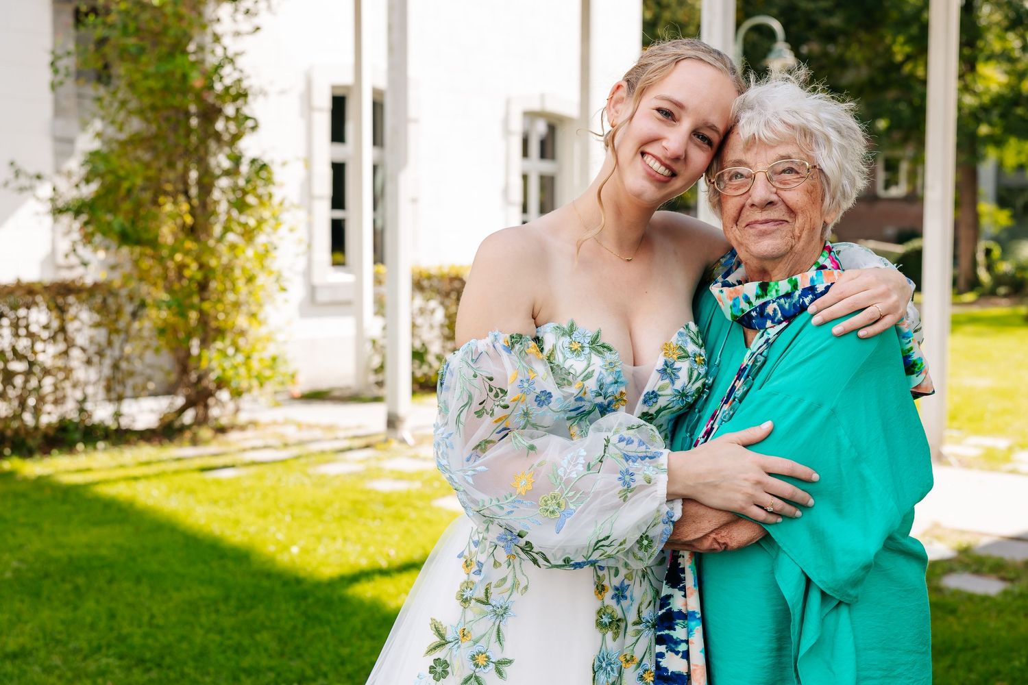 A radiant bride in a floral off-shoulder gown embraces an elderly woman in a sunny garden setting.