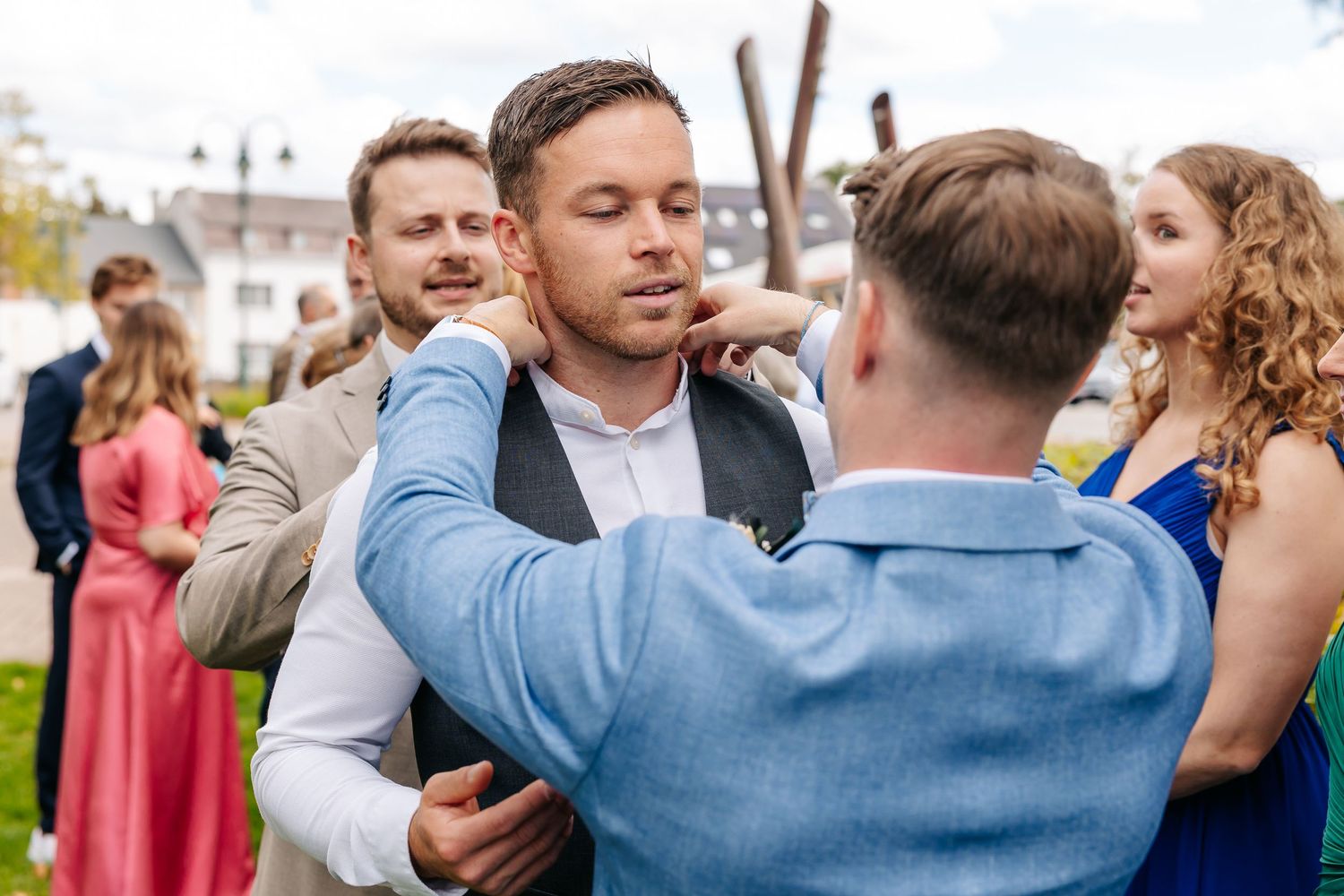Groom in vest adjusts tie with help from groomsman in blue suit at wedding.
