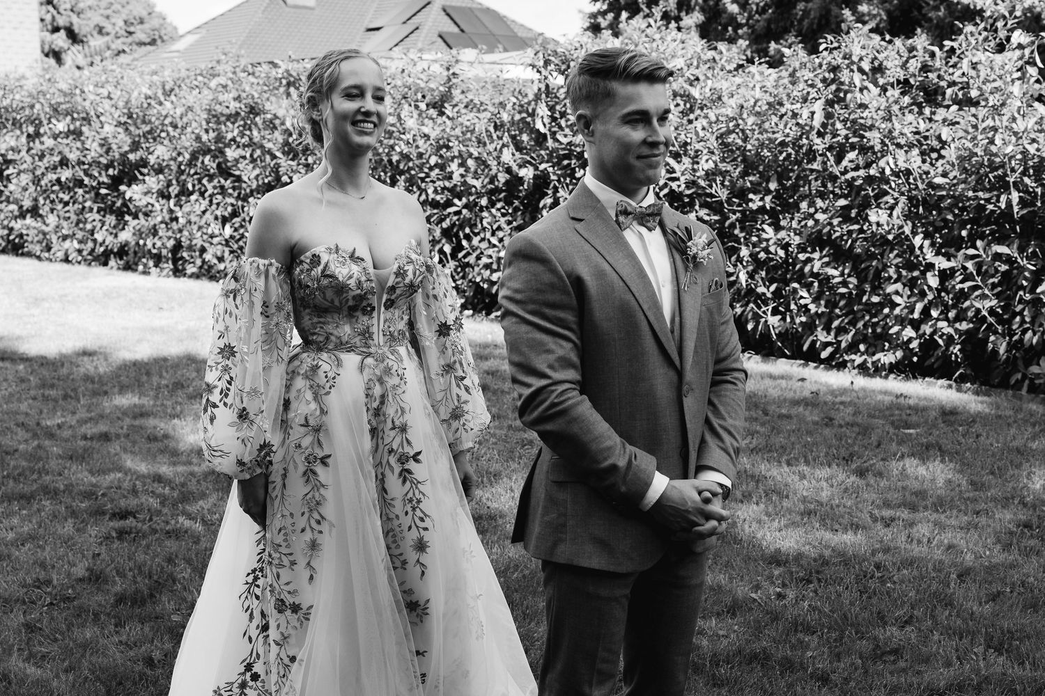 Black and white photo of bride in floral dress and groom in suit sharing a joyful moment outdoors.