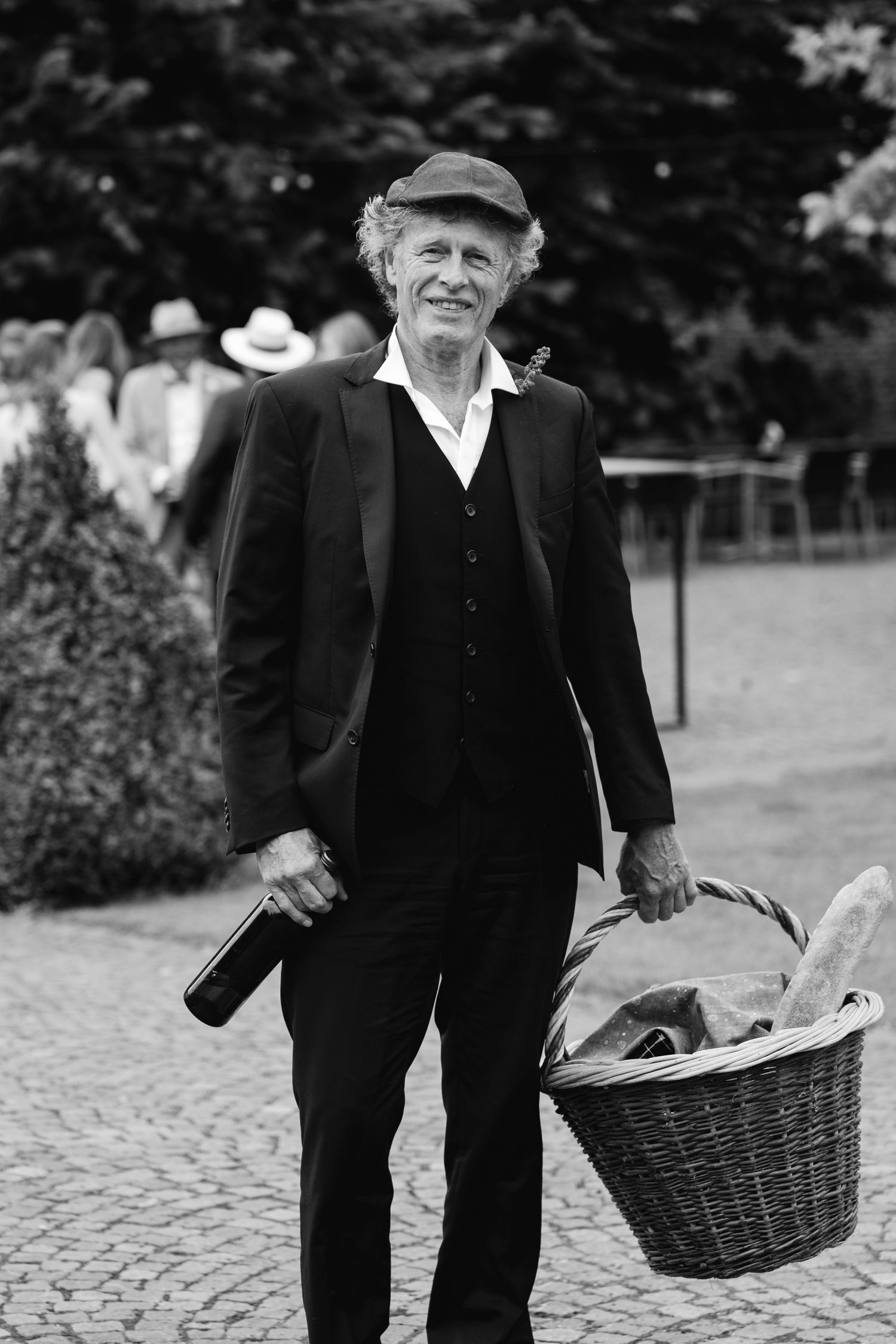 Elderly man in black suit and flat cap carrying a wicker basket and wine bottle at an outdoor wedding.