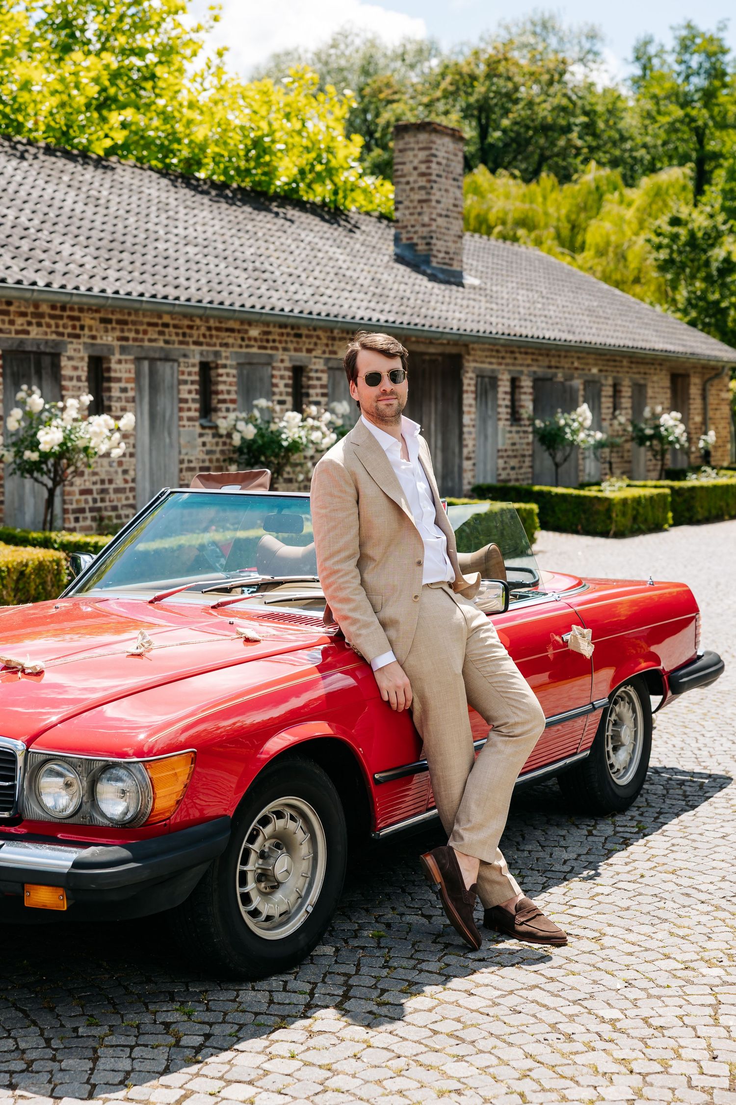 A well-dressed groom in a beige suit leaning against a classic red Mercedes convertible outdoors.