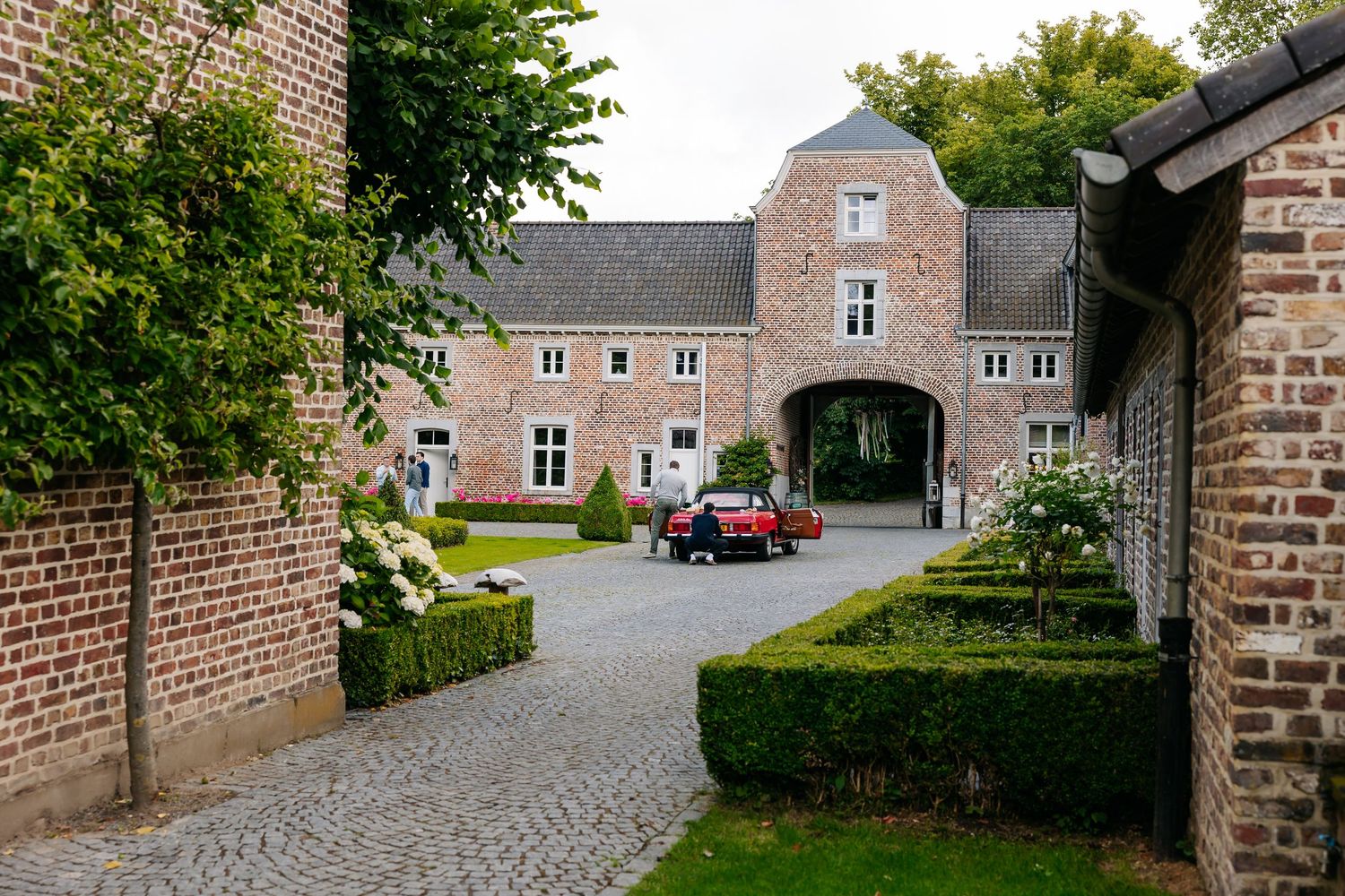 Red classic car parked in courtyard of historic Belgian brick estate.