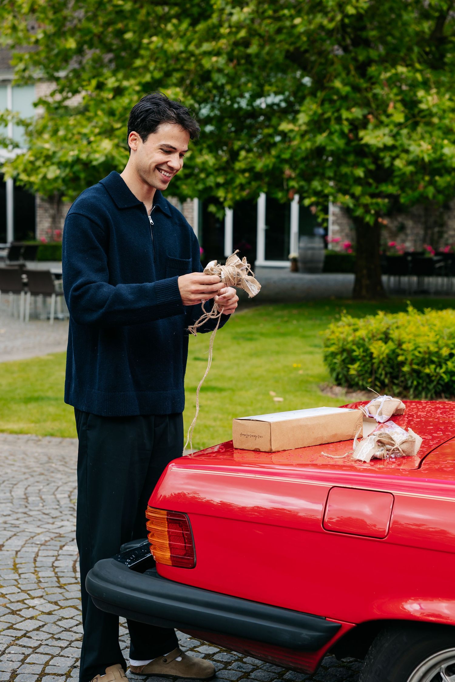 Smiling groom holding wedding shoes next to a red vintage car outdoors.