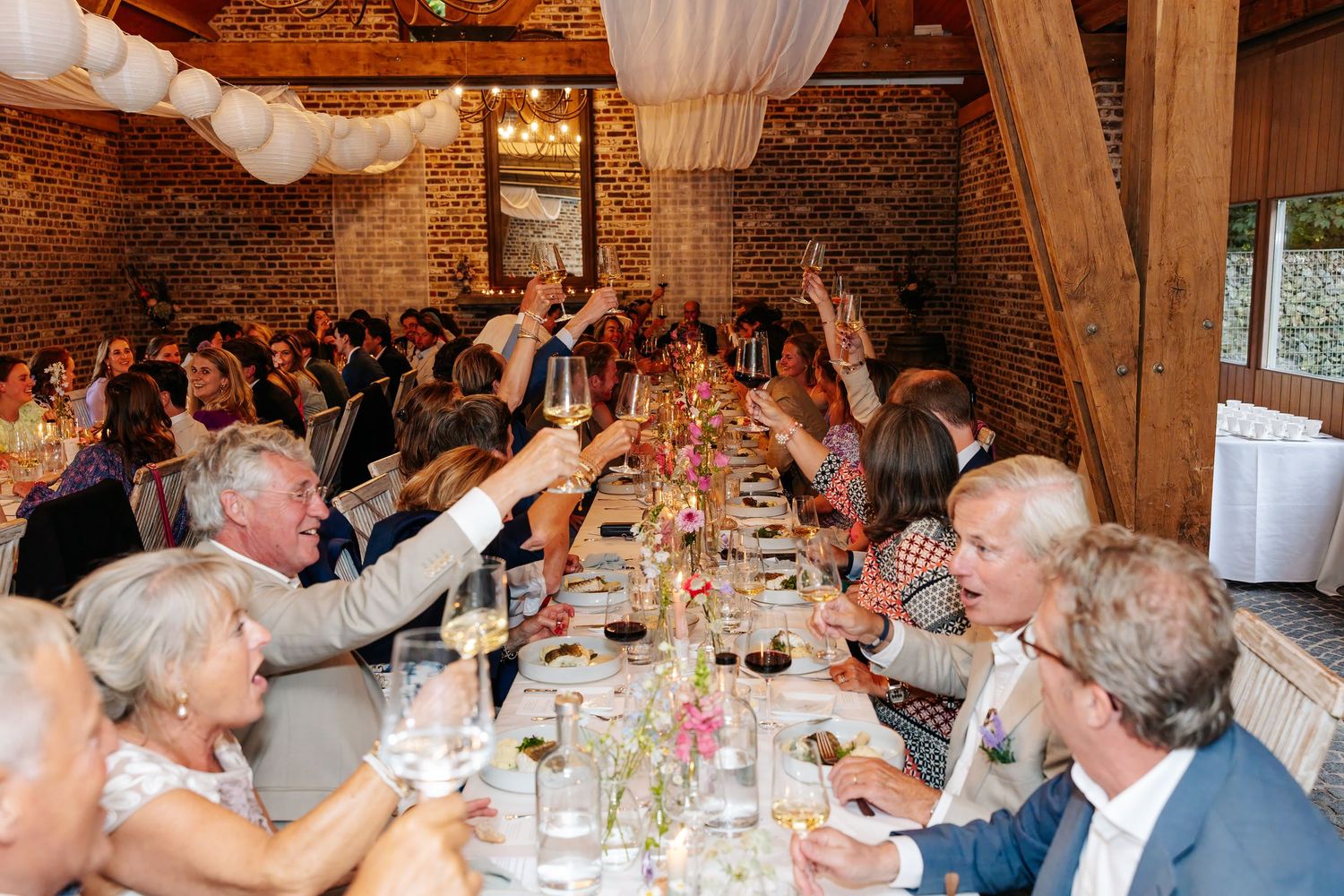 Wedding guests raise glasses in a toast at candlelit reception dinner in a rustic brick barn.
