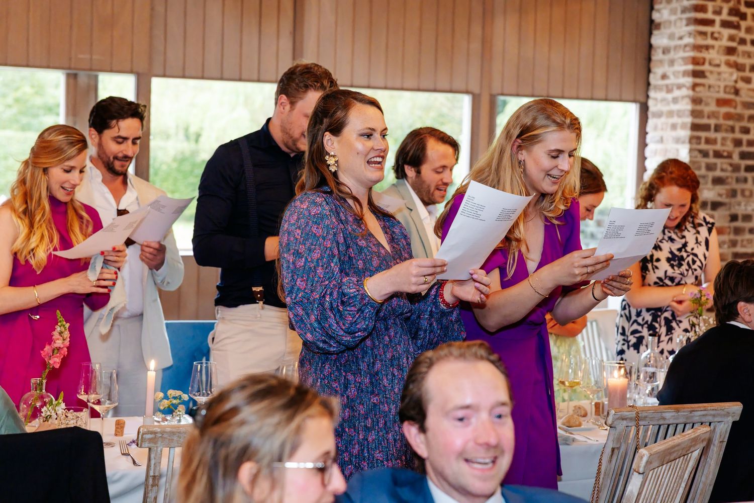 Wedding guests standing and singing from printed sheets at a colorful indoor reception dinner.