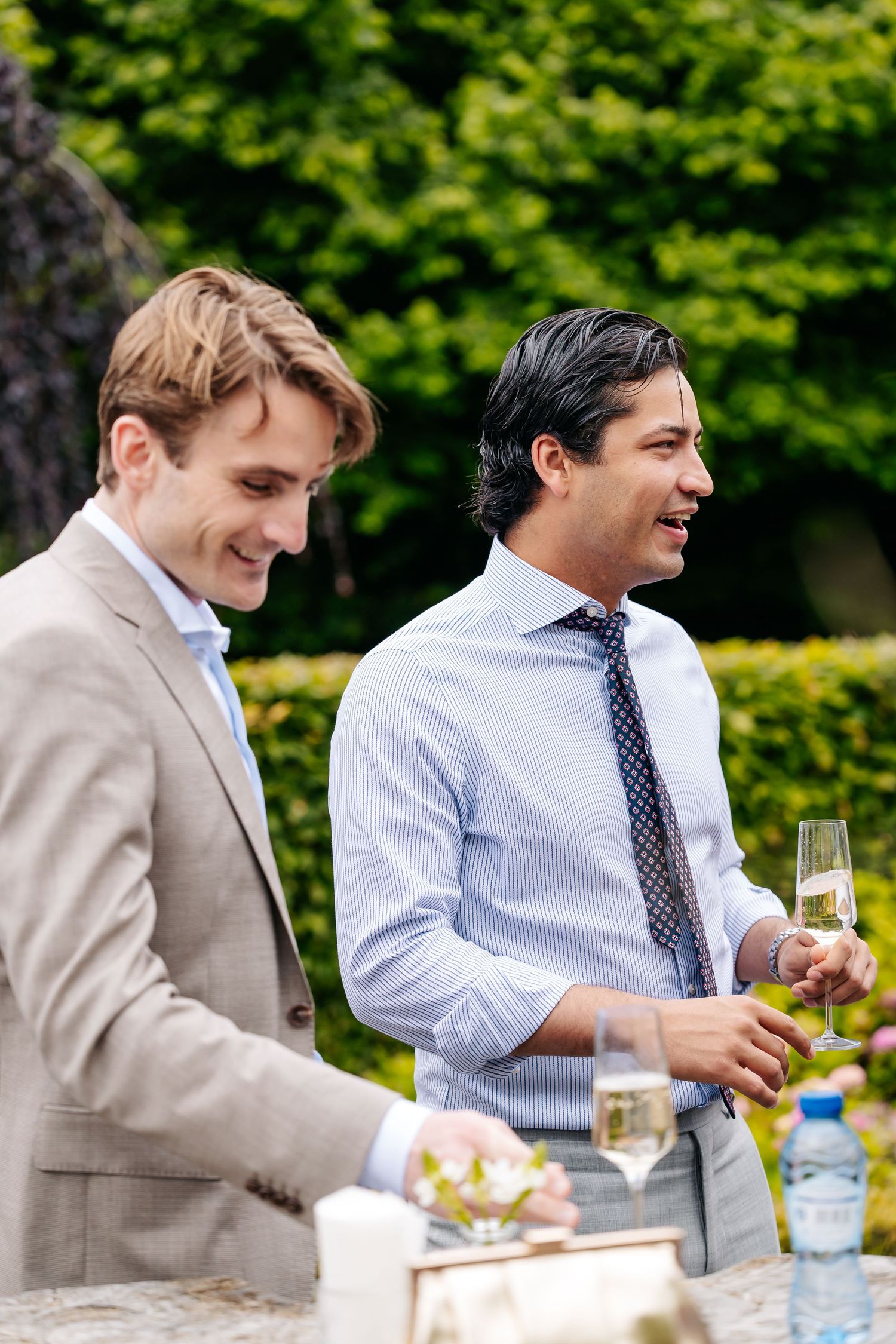 Two well-dressed men smile and enjoy champagne at an outdoor garden wedding reception.